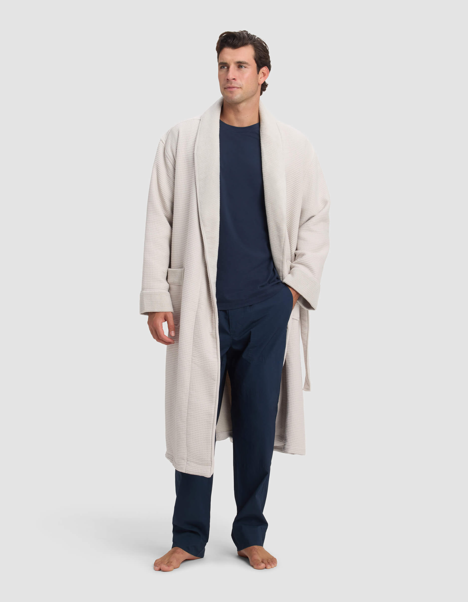 A man stands barefoot in a Cozy Earth Waffle Bath Robe over a navy blue long-sleeve shirt and pants, set against a plain light gray background.|Color:Light Grey
