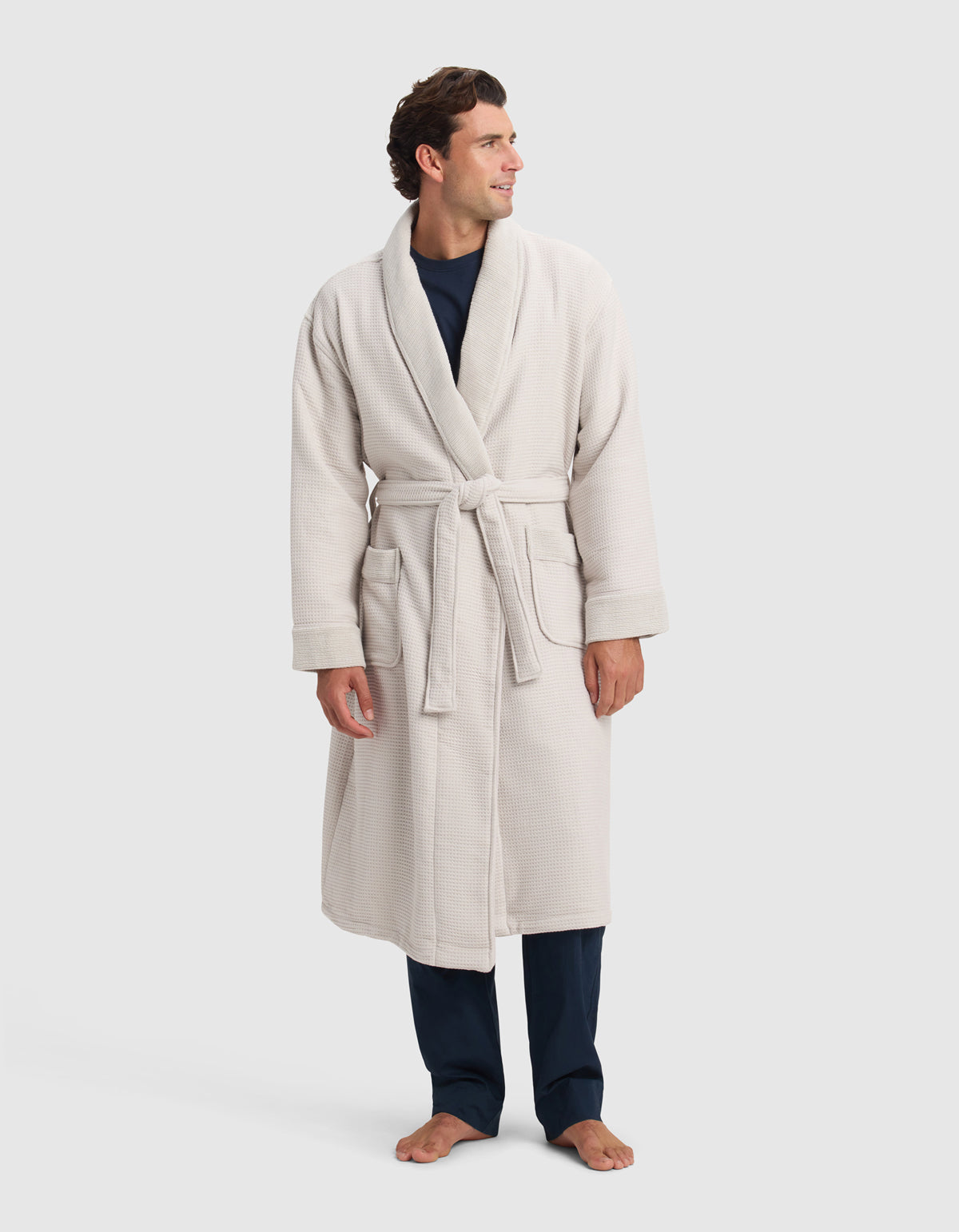 A person wearing a Cozy Earth Waffle Bath Robe, tied at the waist over dark pants and a dark shirt, stands barefoot on a white background, looking off to the side and smiling.