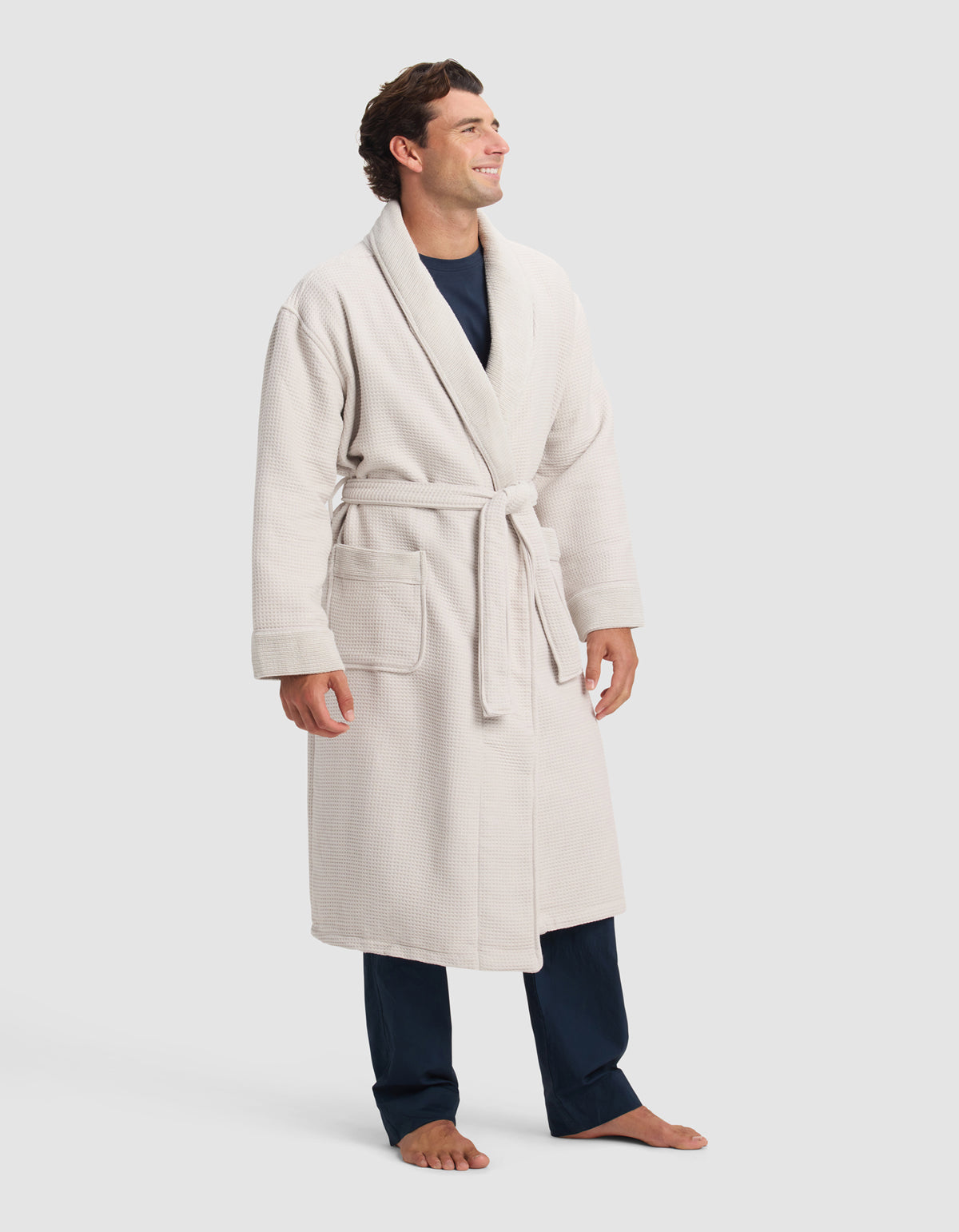 A man stands barefoot, smiling and facing slightly to the side. He is wearing a Cozy Earth Waffle Bath Robe, light-colored and tied at the waist, over dark pajamas, with his hands relaxed by his sides.