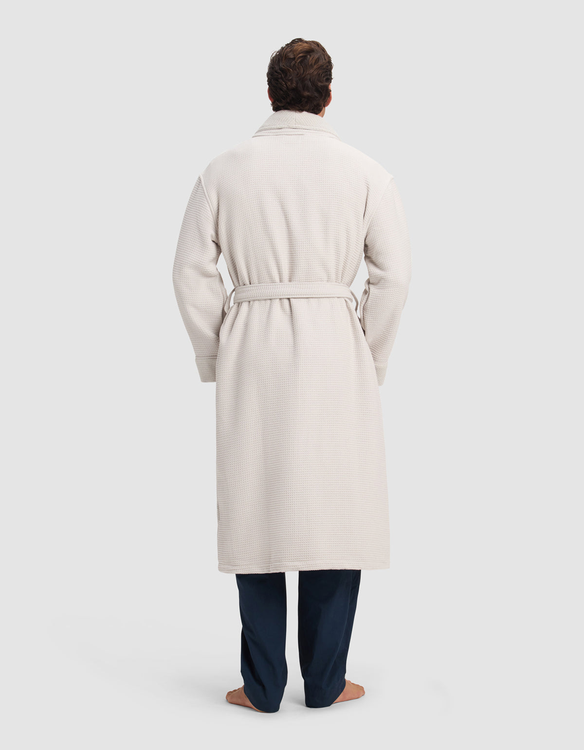 A person stands barefoot facing away against a light background, wearing the Cozy Earth Waffle Bath Robe—a long, light-colored robe with a belt—over dark pants.