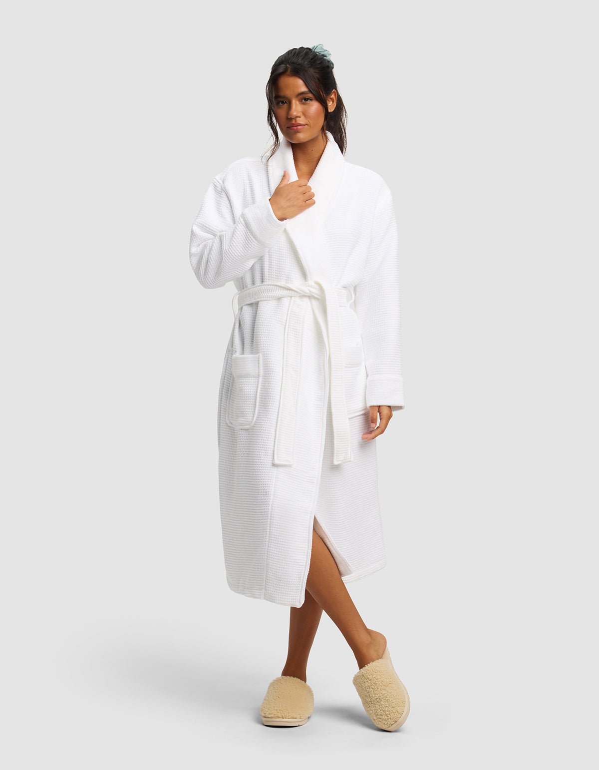 A woman stands against a plain background wearing the Cozy Earth Waffle Bath Robe tied at the waist and beige slippers. Her hair is tied back, with one hand near her collar and one at her side.