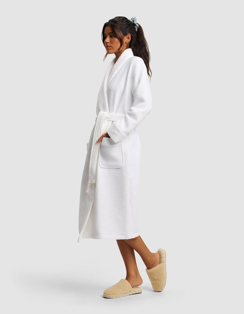 A woman in profile wears the Cozy Earth Waffle Bath Robe—white, with front pockets and a waist tie—and beige slippers. Her long dark hair is pulled back with a light blue scrunchie against a plain light gray background.