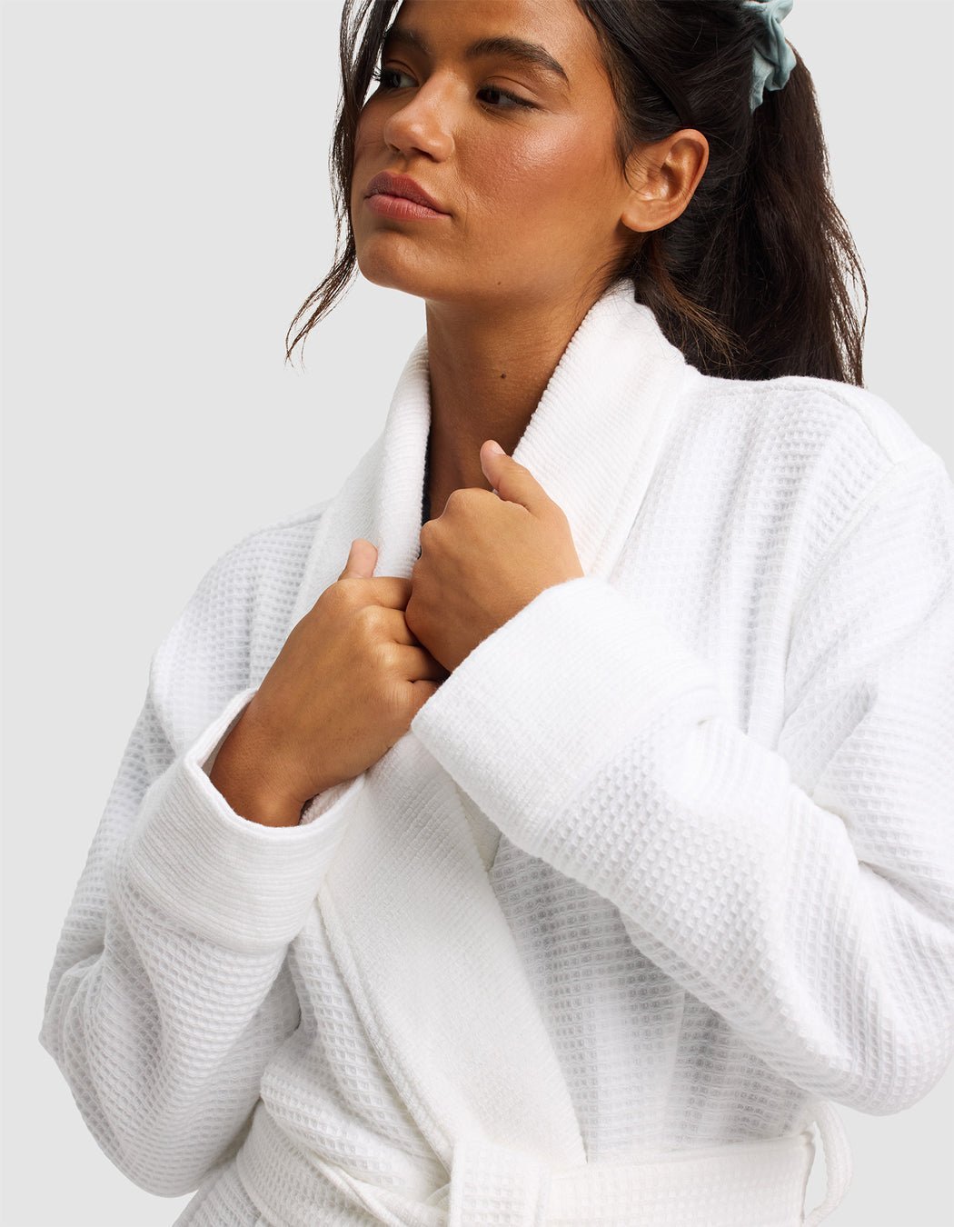 A woman with a blue scrunchie wears the Cozy Earth Waffle Bath Robe, holding the collar with both hands and gazing to the side against a plain light background.