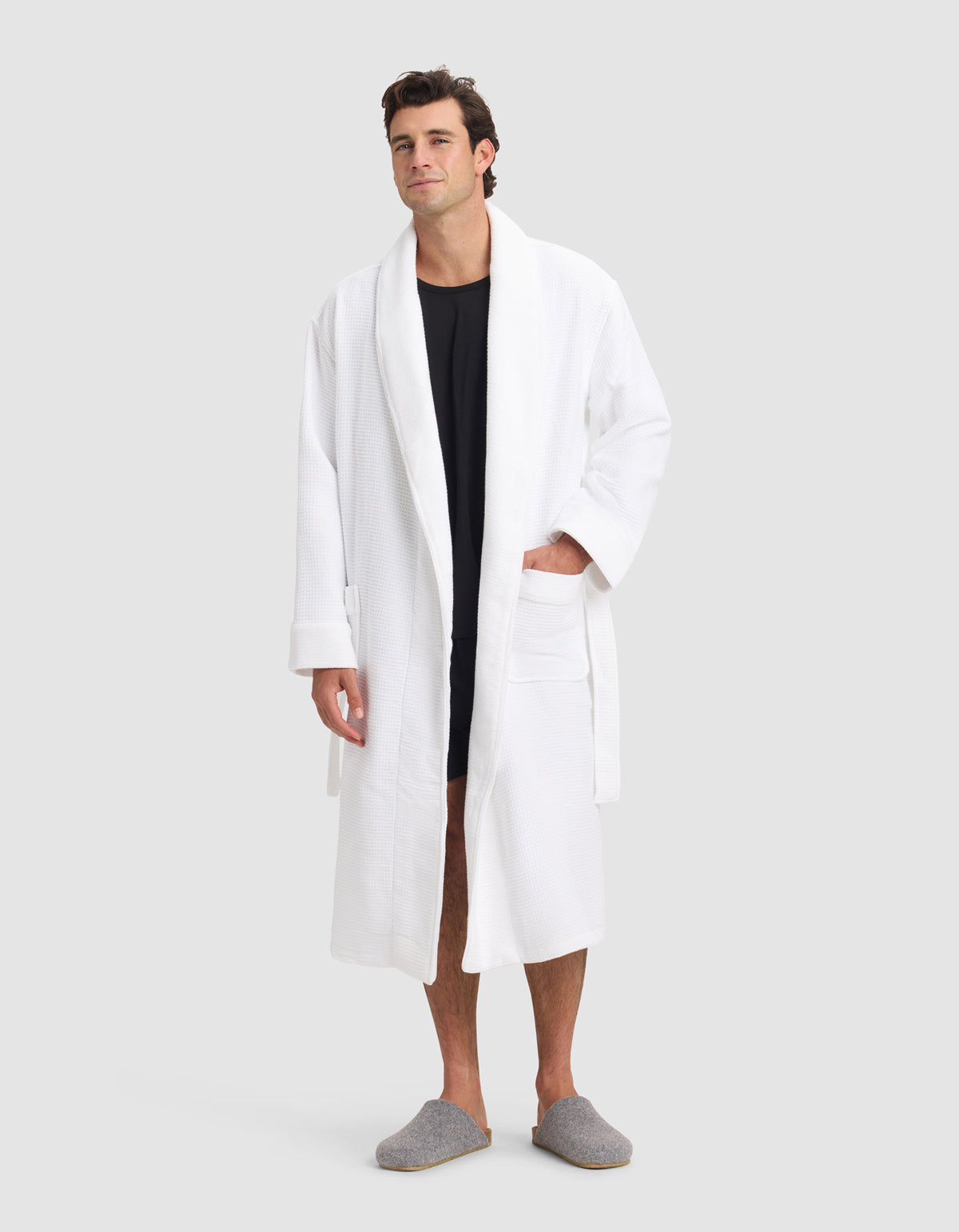 A man stands facing forward against a plain light grey background, wearing the Cozy Earth Waffle Bath Robe over a black shirt with grey slippers. One hand rests in the robe’s pocket.