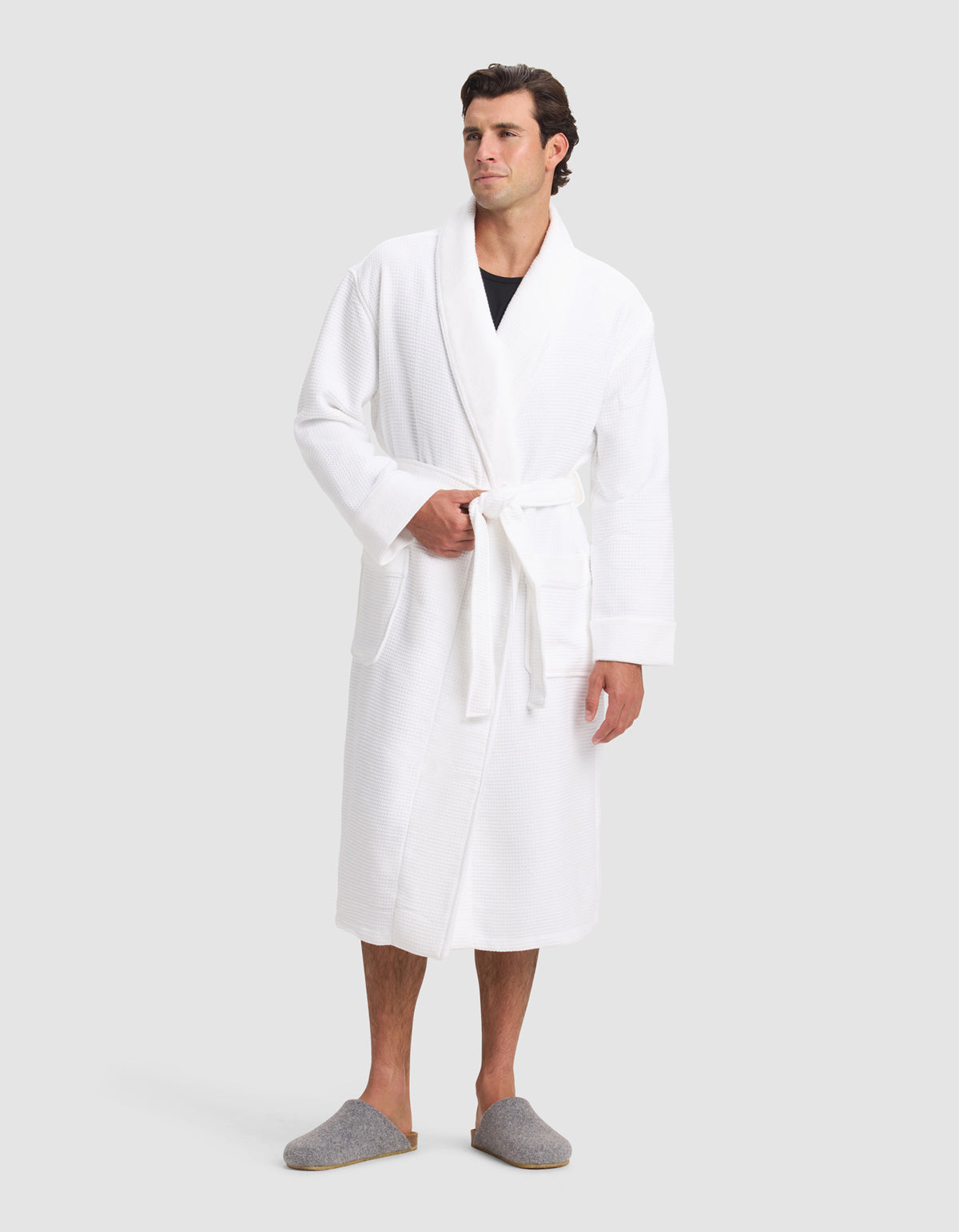 A man wears the Cozy Earth Waffle Bath Robe with a tied belt over a dark shirt, paired with gray slippers, standing against a plain light gray background.