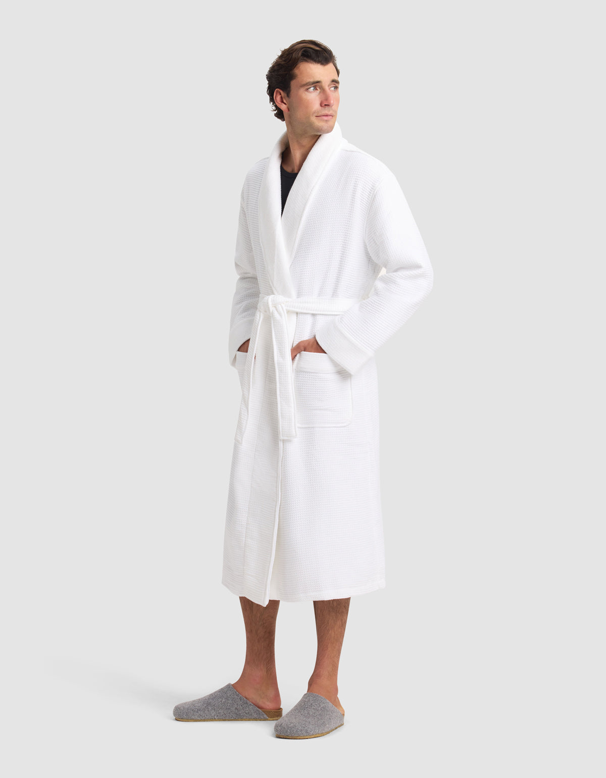 A man stands against a light gray background, wearing the Cozy Earth Waffle Bath Robe with a belted waist and pockets, along with gray slippers. He looks slightly to the side.