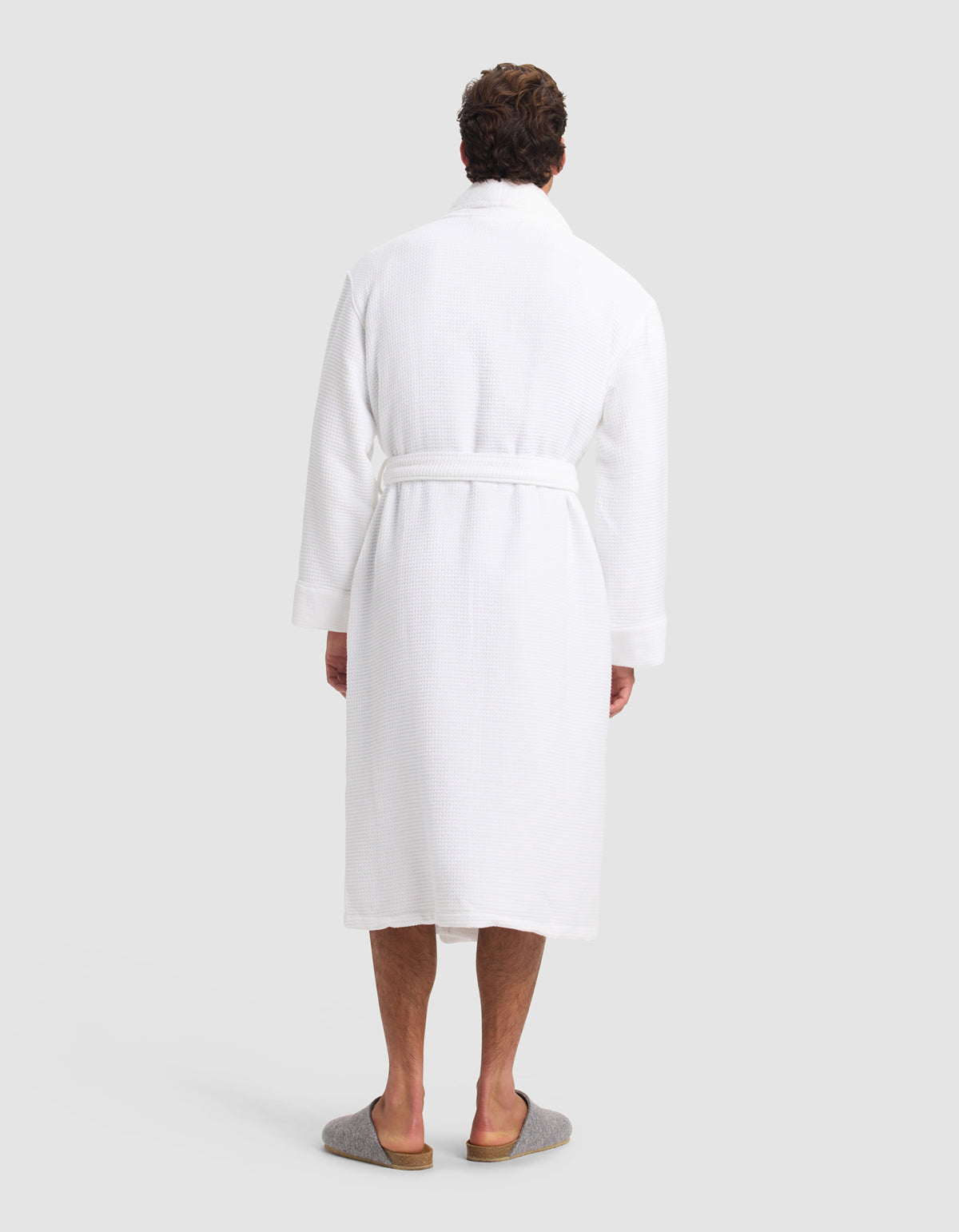 A person faces away, wearing the Cozy Earth Waffle Bath Robe—a white, knee-length robe with a belt—paired with gray slippers against a plain light background.