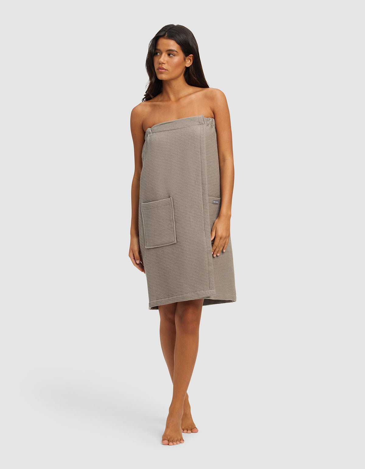 A woman stands barefoot against a plain background, wearing the Cozy Earth Waffle Bath Wrap in grey, featuring a waffle texture, front pocket, and chest-to-knee coverage.|Color:Charcoal