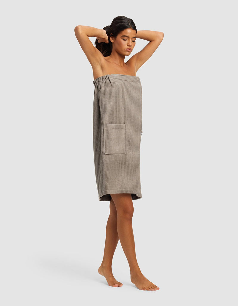 A woman with medium skin tone stands barefoot against a light gray background, adjusting her hair while wearing the Cozy Earth Waffle Bath Wrap in taupe, knee-length style with a front pocket.