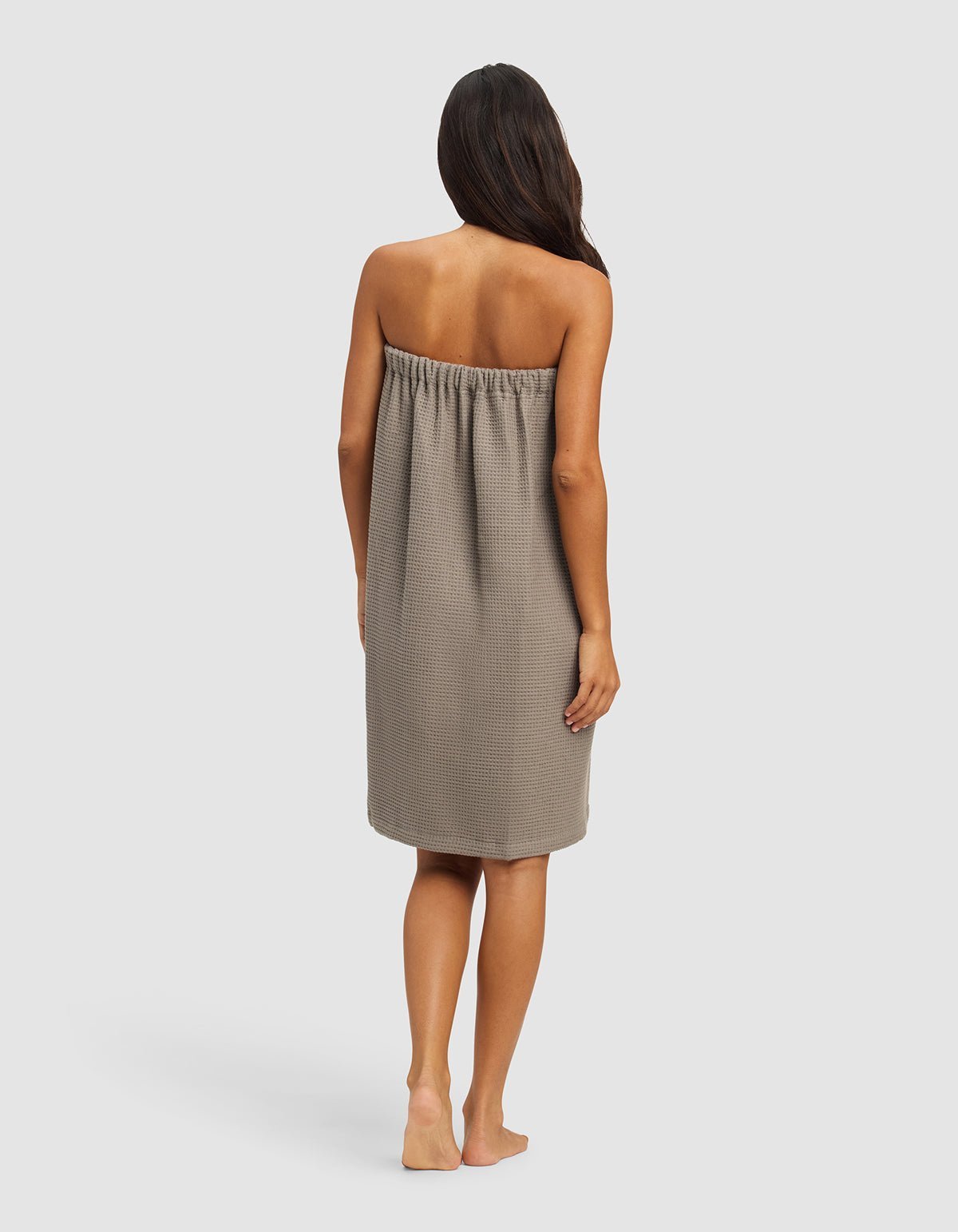 A woman with long, dark hair stands barefoot on a plain background, facing away from the camera. She wears the Cozy Earth Waffle Bath Wrap, a strapless, knee-length towel wrap in taupe with a textured pattern.|Color:Charcoal