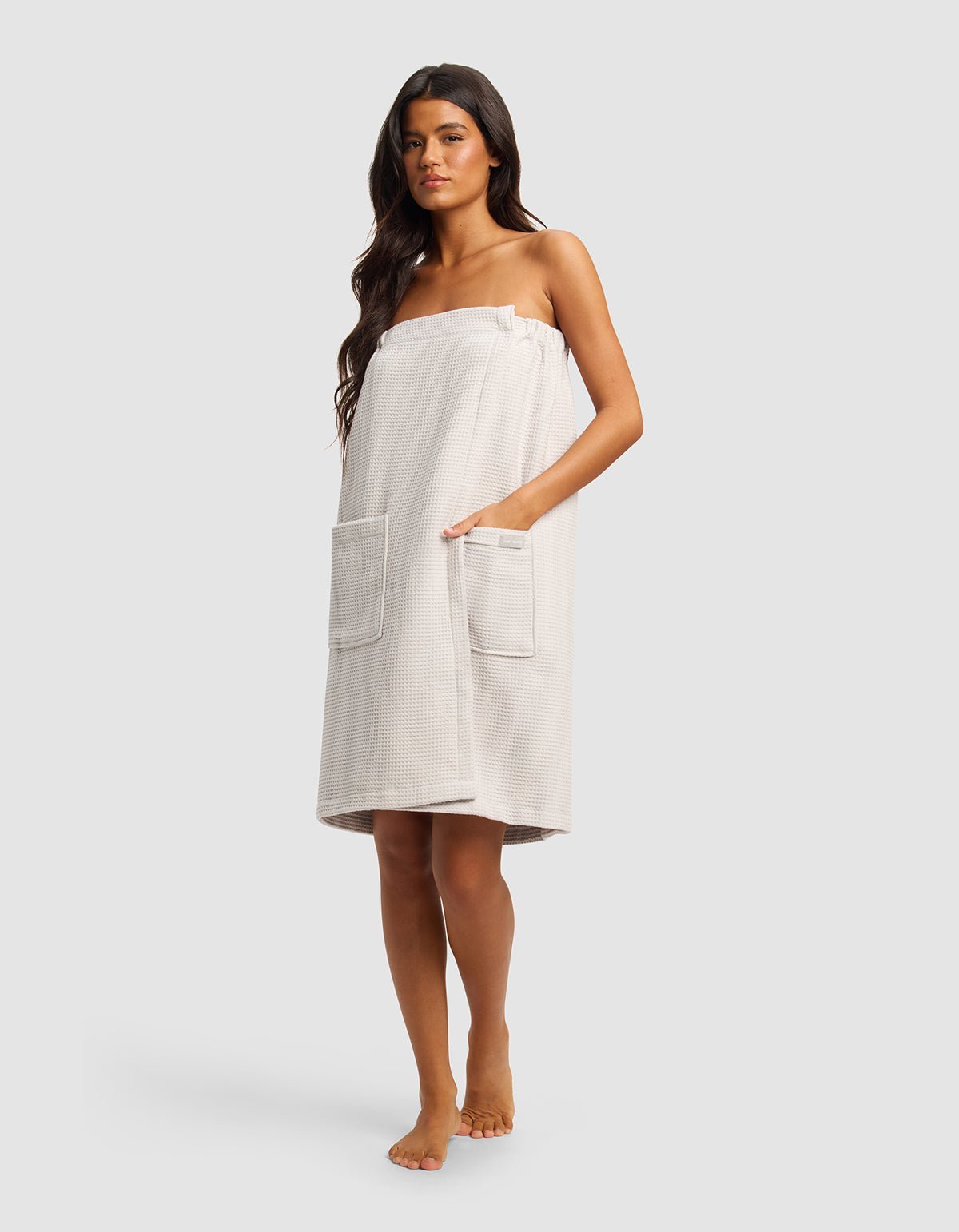 A woman with long dark hair stands barefoot wearing the Cozy Earth Waffle Bath Wrap in light beige, featuring two large front pockets and covering her from chest to knees. The background is plain and light gray.|Color:Light Grey