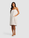 A woman with long dark hair stands barefoot wearing the Cozy Earth Waffle Bath Wrap in light beige, featuring two large front pockets and covering her from chest to knees. The background is plain and light gray.|Color:Light Grey