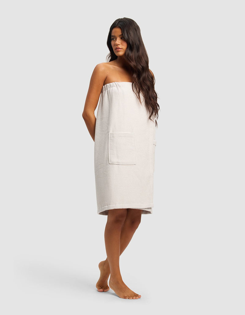 A woman with long dark hair stands barefoot, wearing the Cozy Earth Waffle Bath Wrap—a strapless, light towel wrap with large front pockets—facing slightly to the side against a plain, light background.