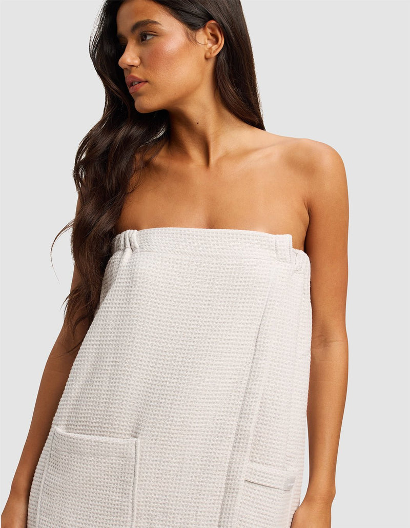 A woman with long, dark hair wears the Cozy Earth Waffle Bath Wrap, featuring pockets and a textured light fabric, standing against a plain light background and looking to the side.
