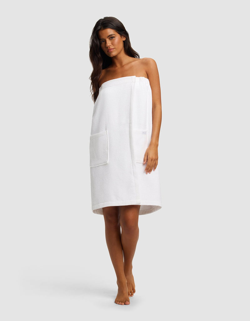 A woman with long dark hair stands barefoot, wearing the Cozy Earth Waffle Bath Wrap with a large front pocket. The towel covers her chest to above her knees as she poses against a plain light gray background.
