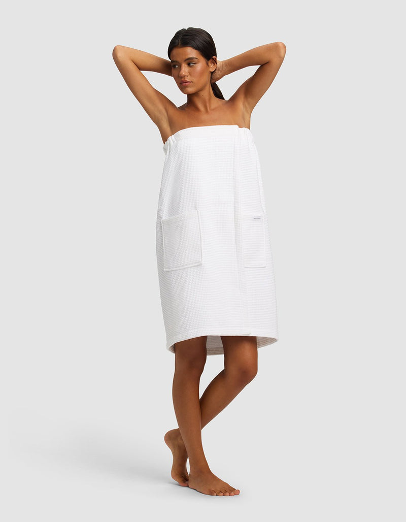A woman stands barefoot with her arms raised, wearing the Cozy Earth Waffle Bath Wrap with large front pockets against a plain light gray background.