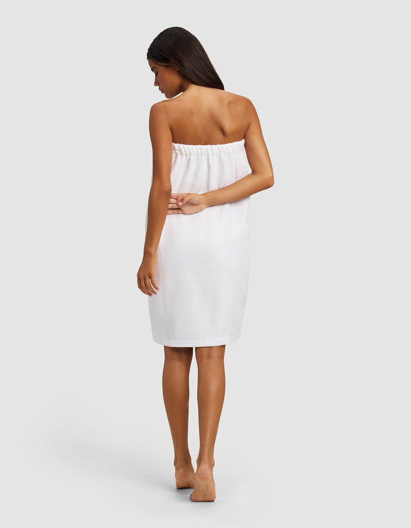 A woman with long hair stands barefoot on a light background, wearing the white, knee-length Cozy Earth Waffle Bath Wrap. Seen from behind, her left arm rests behind her back as she looks slightly to the side.