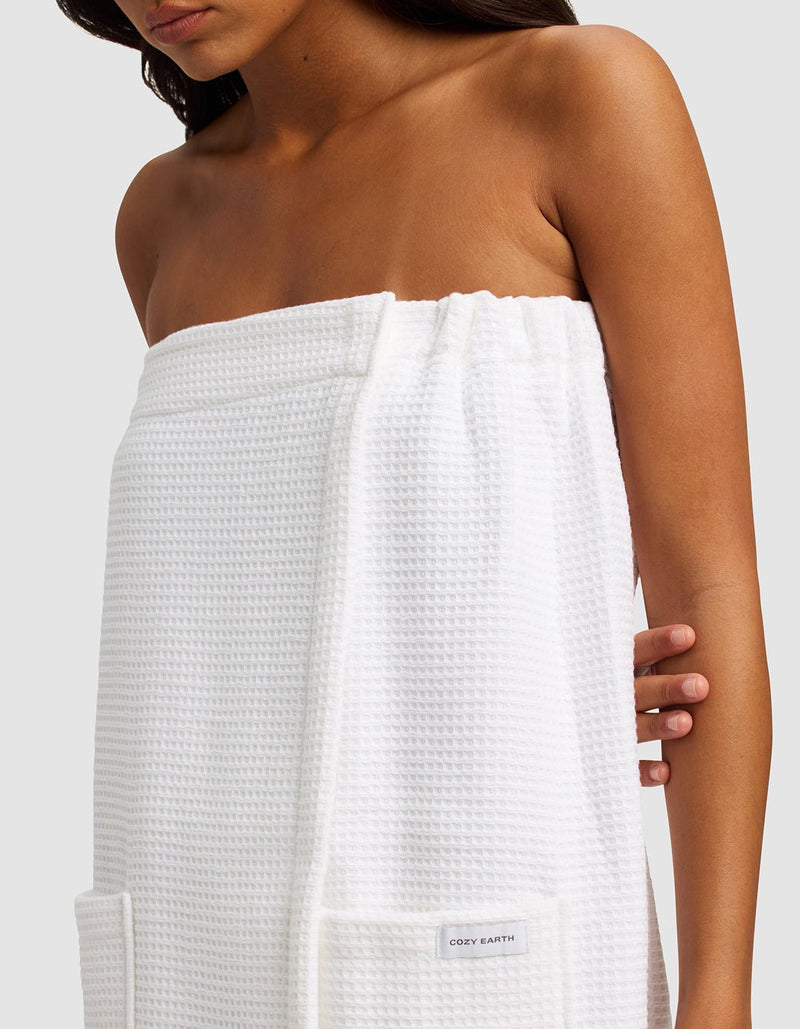 A woman with long dark hair is wrapped in the Cozy Earth Waffle Bath Wrap, a white, textured towel with pockets. She stands against a plain light gray background, shown from shoulders to mid-thigh.