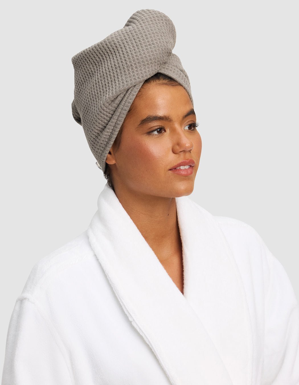 A person wears a white bathrobe and a Cozy Earth Waffle Hair Towel in light gray, wrapped around their head, looking slightly to the side with a neutral expression against a plain light gray background. |Color:Charcoal