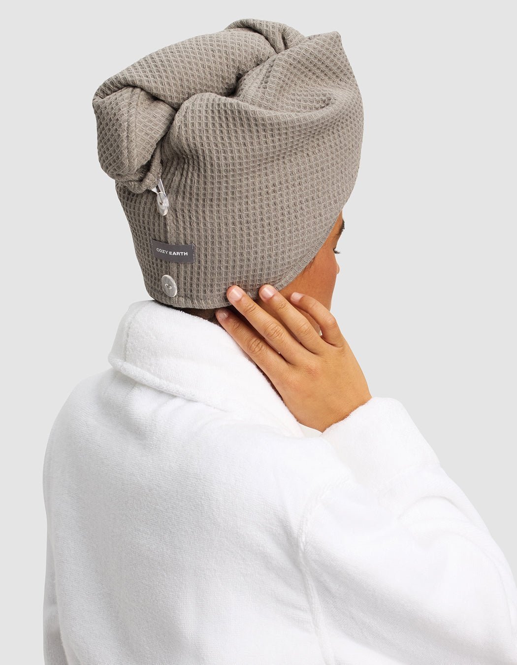 A person is shown from behind wearing the Cozy Earth Waffle Hair Towel in taupe and a white bathrobe, with one hand touching their neck, against a plain background.