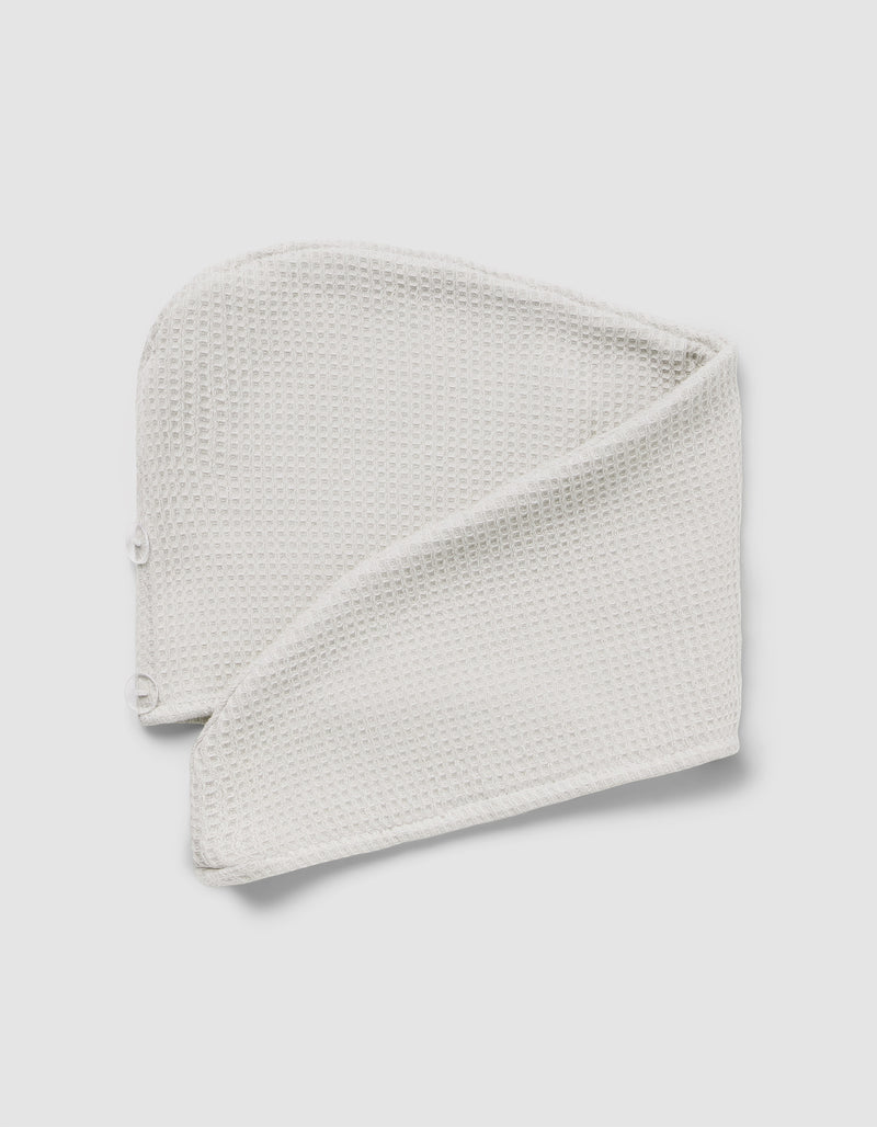The Cozy Earth Waffle Hair Towel, a folded, textured white headband with snap buttons at one end, is displayed on a light gray background.