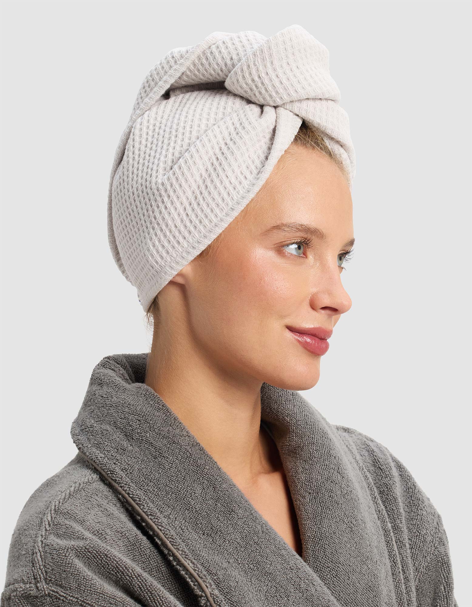 A woman in a gray bathrobe, facing right and smiling slightly, wears the Cozy Earth Waffle Hair Towel in light beige wrapped around her head. Her hair is covered, and the background is plain light gray. 