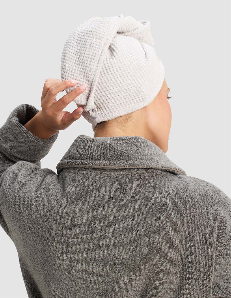 A person in a gray bathrobe is seen from behind, adjusting a Cozy Earth Waffle Hair Towel wrapped around their head. The background is plain and light-colored.