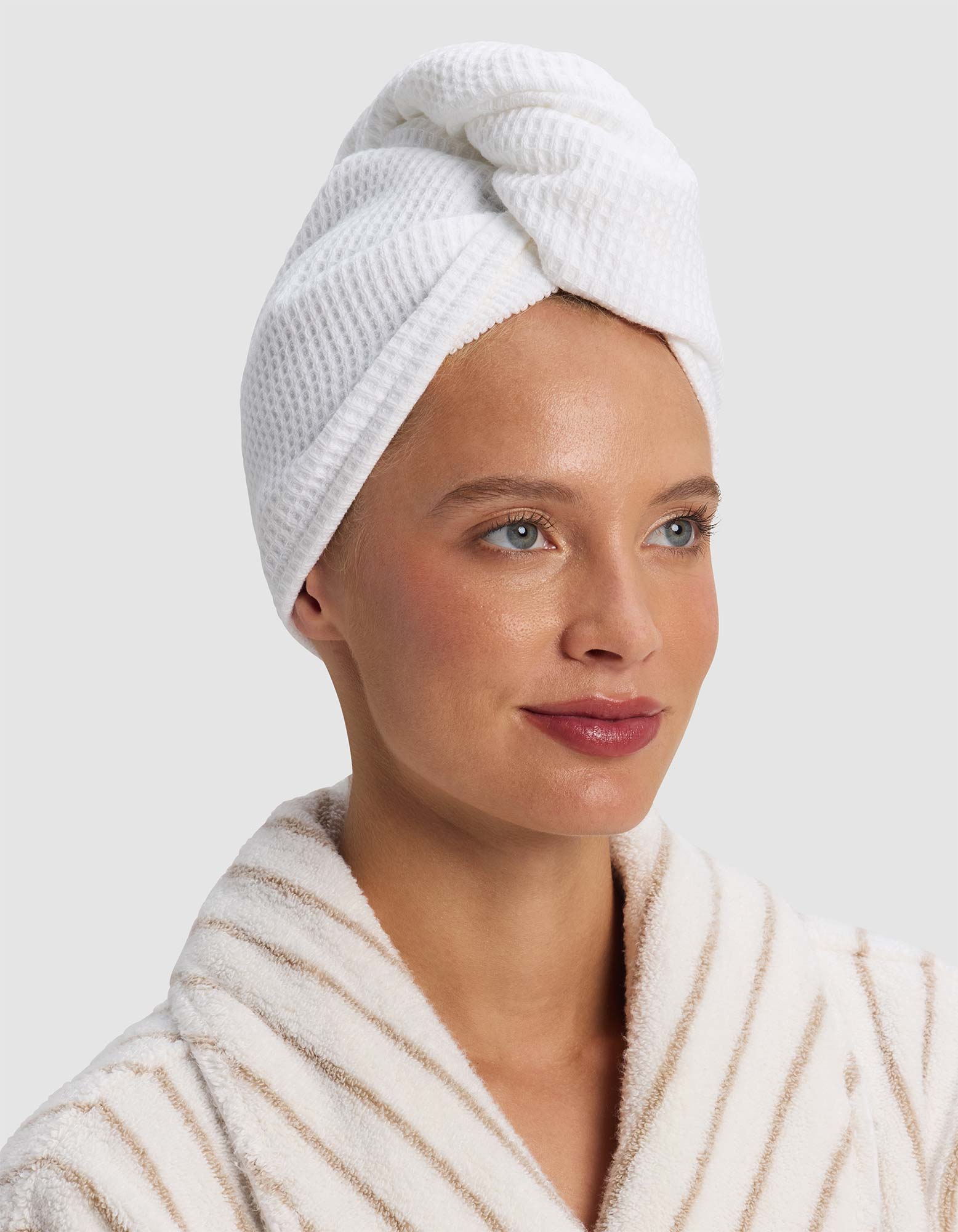 A person wearing the Cozy Earth Waffle Hair Towel and a cream-and-beige striped bathrobe looks slightly to the side with a gentle smile.|Color:White