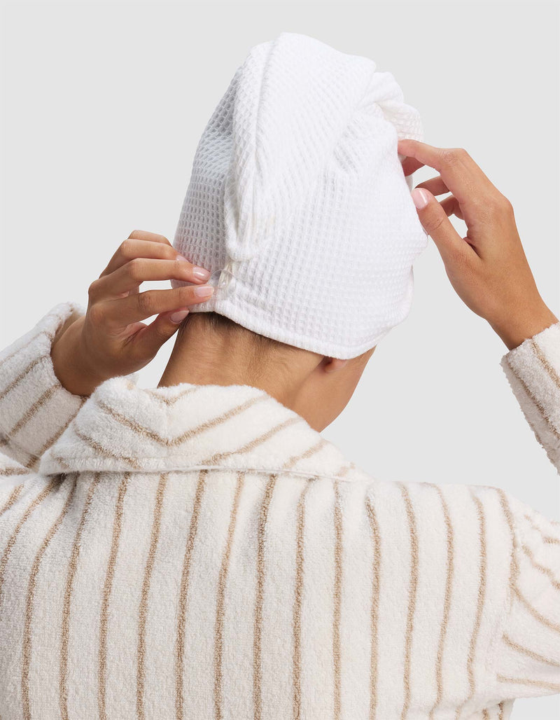 A person shown from the back, against a plain light background, wears a striped bathrobe and adjusts a Cozy Earth Waffle Hair Towel wrapped around their head.
