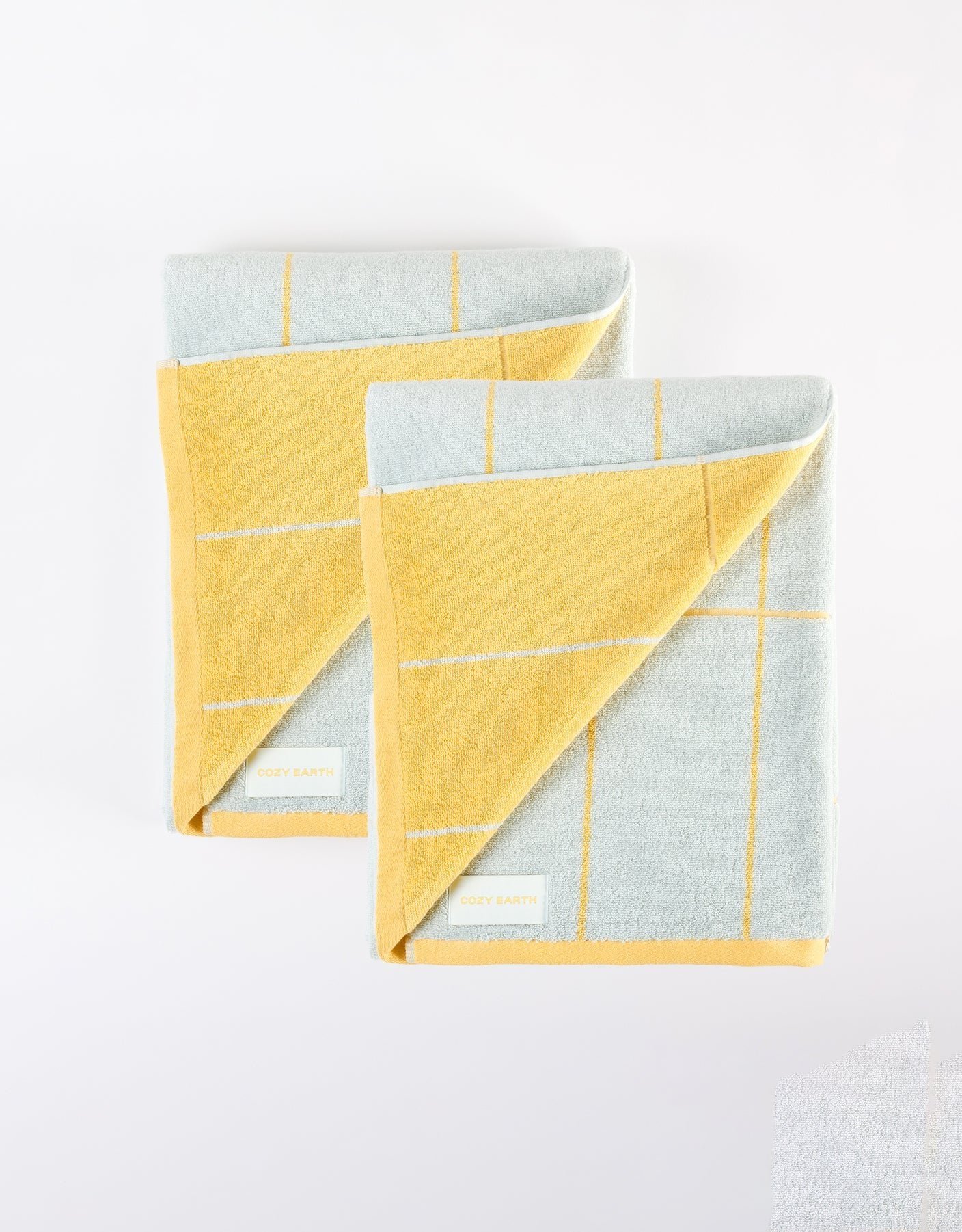The Windowpane Resort Towel Bundle by Cozy Earth features two folded towels with a light blue and yellow checkered pattern, yellow edges, and a white "COZY EARTH" label. Both are displayed on a white surface. |Color:Sunny Sky