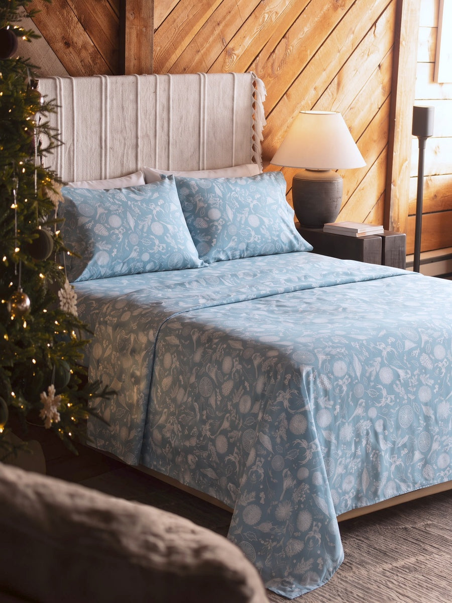 A bed dressed in a light blue and white floral patterned comforter and matching pillow shams, set in a rustic bedroom with wood-paneled walls, a bedside lamp, and a Christmas tree at the side.