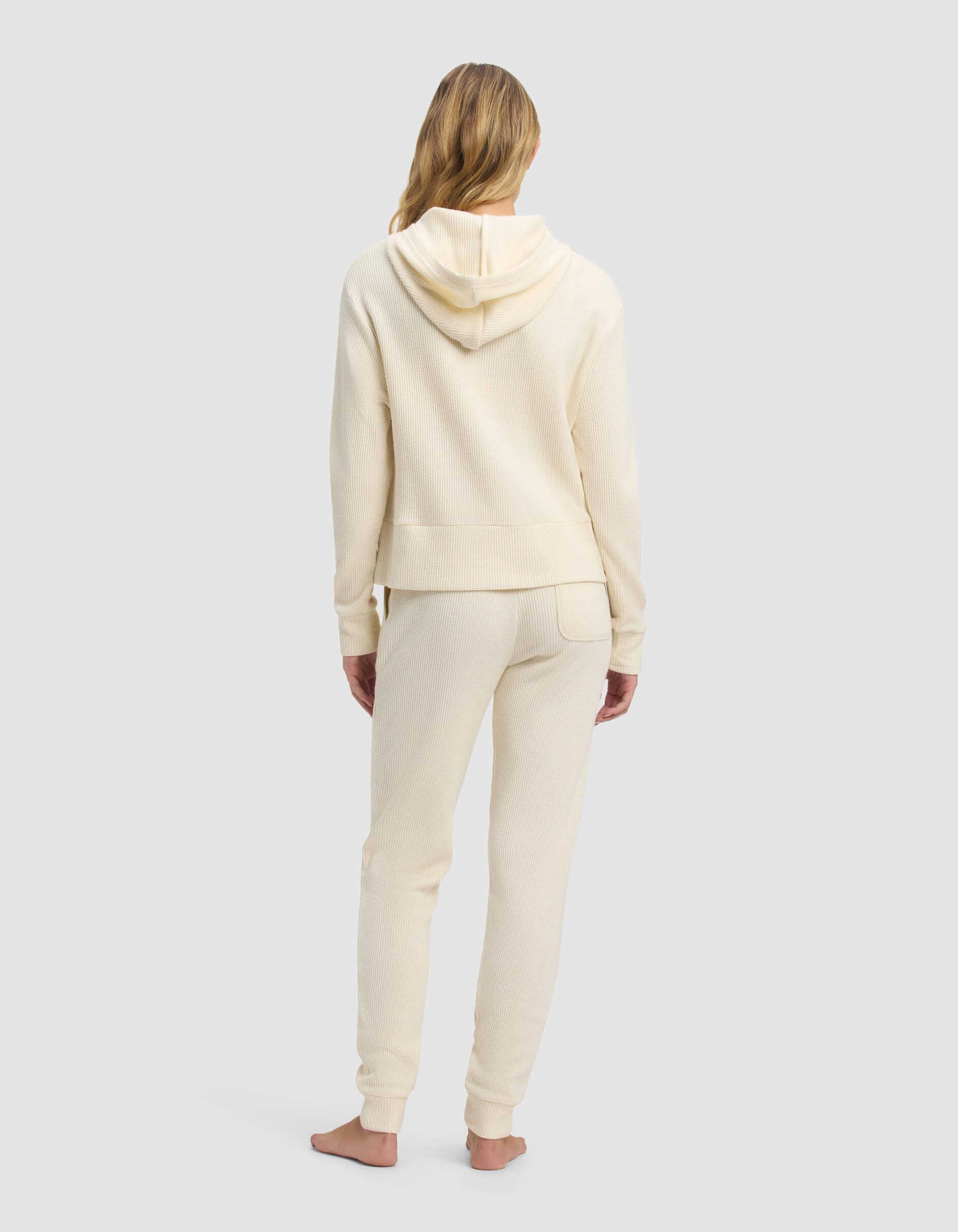 A woman with long blonde hair stands barefoot, facing away, wearing the Cozy Earth Women's Blake Waffle Hoodie & Jogger Set in cream against a plain light gray background.