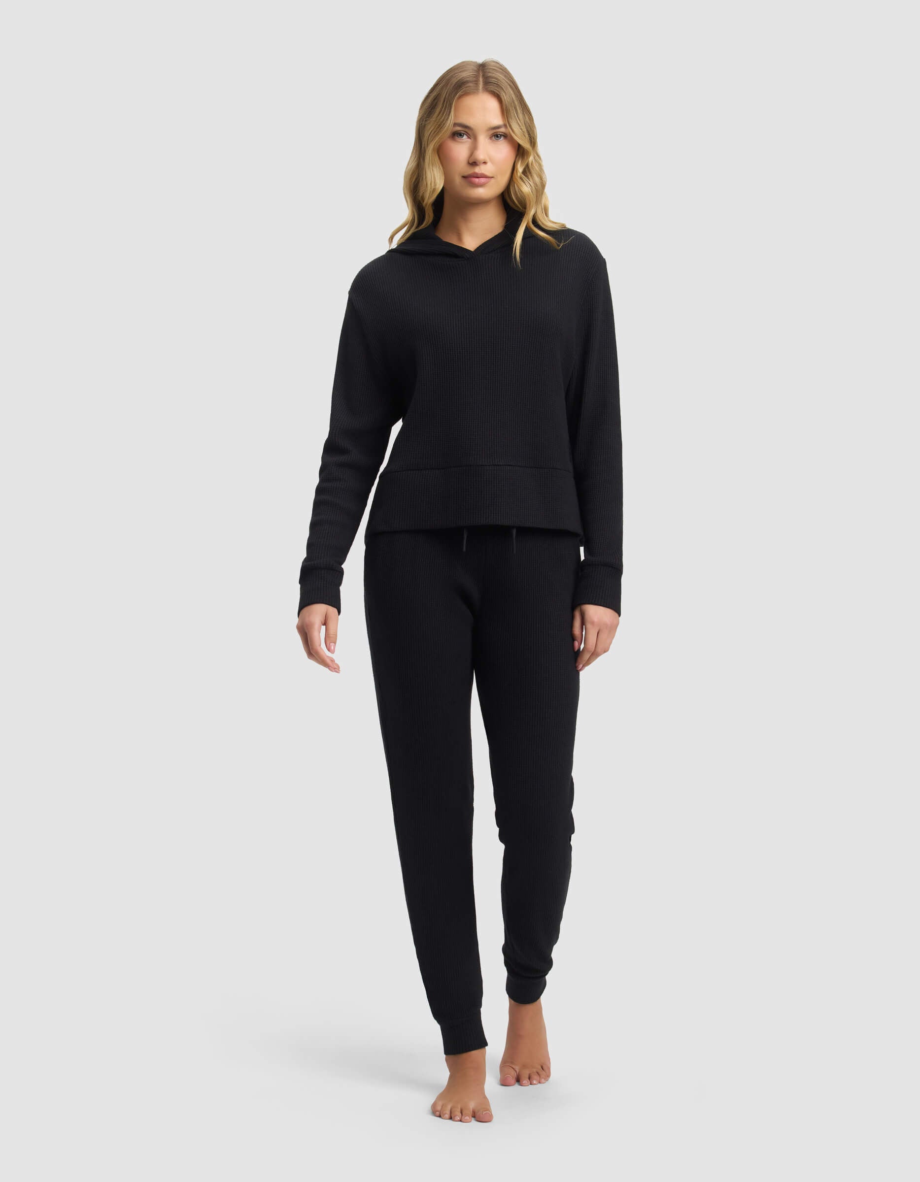 A woman with blonde hair models the Cozy Earth Women's Blake Waffle Hoodie & Jogger Set in black, standing barefoot against a plain light gray background.