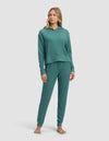 A woman stands barefoot in the Cozy Earth Women's Blake Waffle Hoodie & Jogger Set, featuring a matching teal long-sleeve hoodie and drawstring pants, against a plain light background.