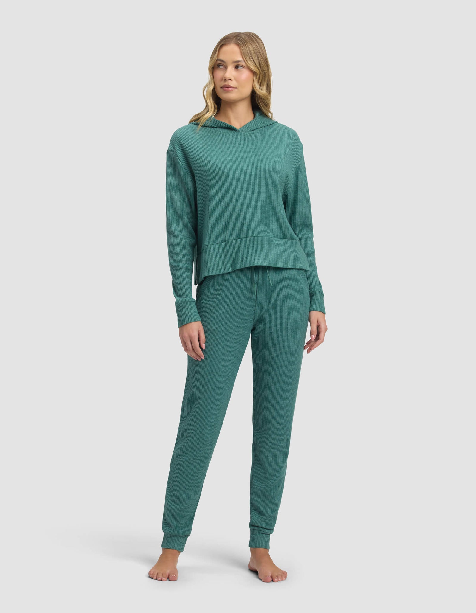 A woman stands barefoot in the Cozy Earth Women's Blake Waffle Hoodie & Jogger Set, featuring a matching teal long-sleeve hoodie and drawstring pants, against a plain light background.