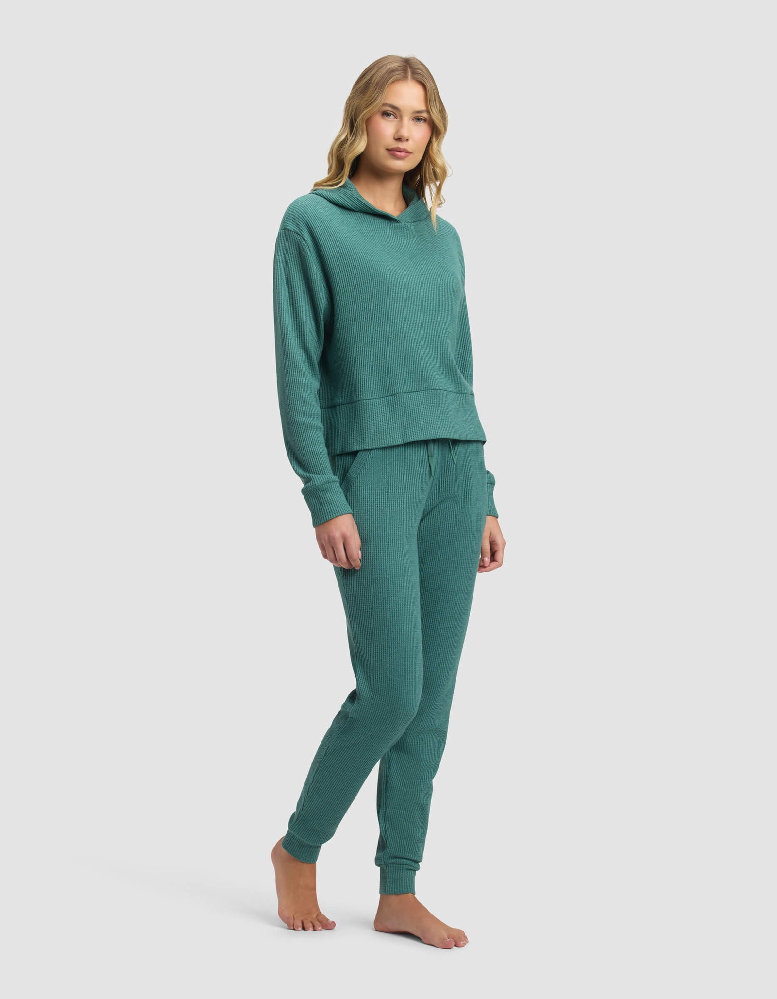 A woman with wavy blonde hair stands barefoot against a plain background, wearing the Cozy Earth Women's Blake Waffle Hoodie & Jogger Set in teal, featuring a long-sleeve hoodie and jogger pants, looking relaxed.
