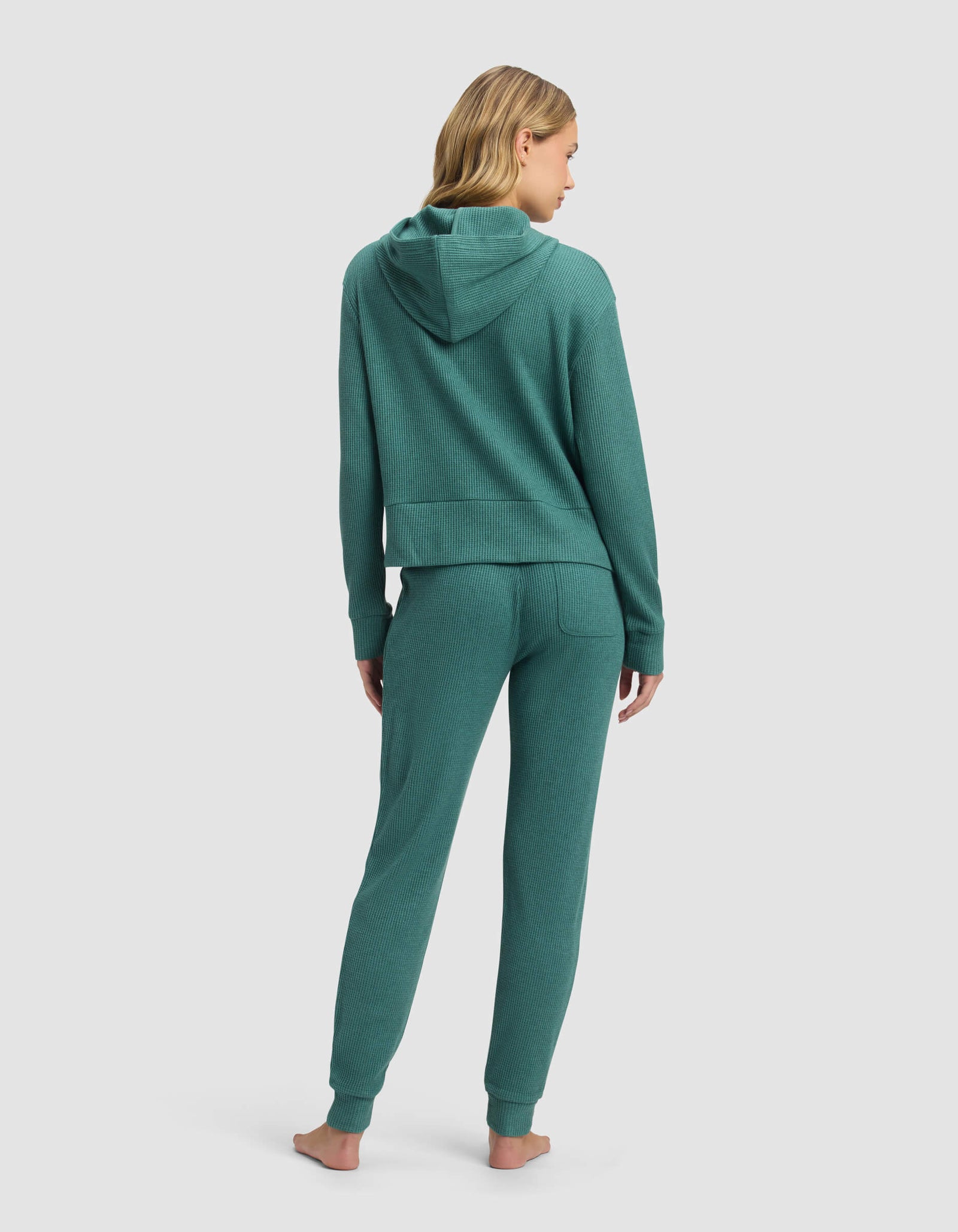 A woman with long blonde hair stands barefoot, facing away, wearing the Cozy Earth Women's Blake Waffle Hoodie & Jogger Set in teal against a plain light gray background.