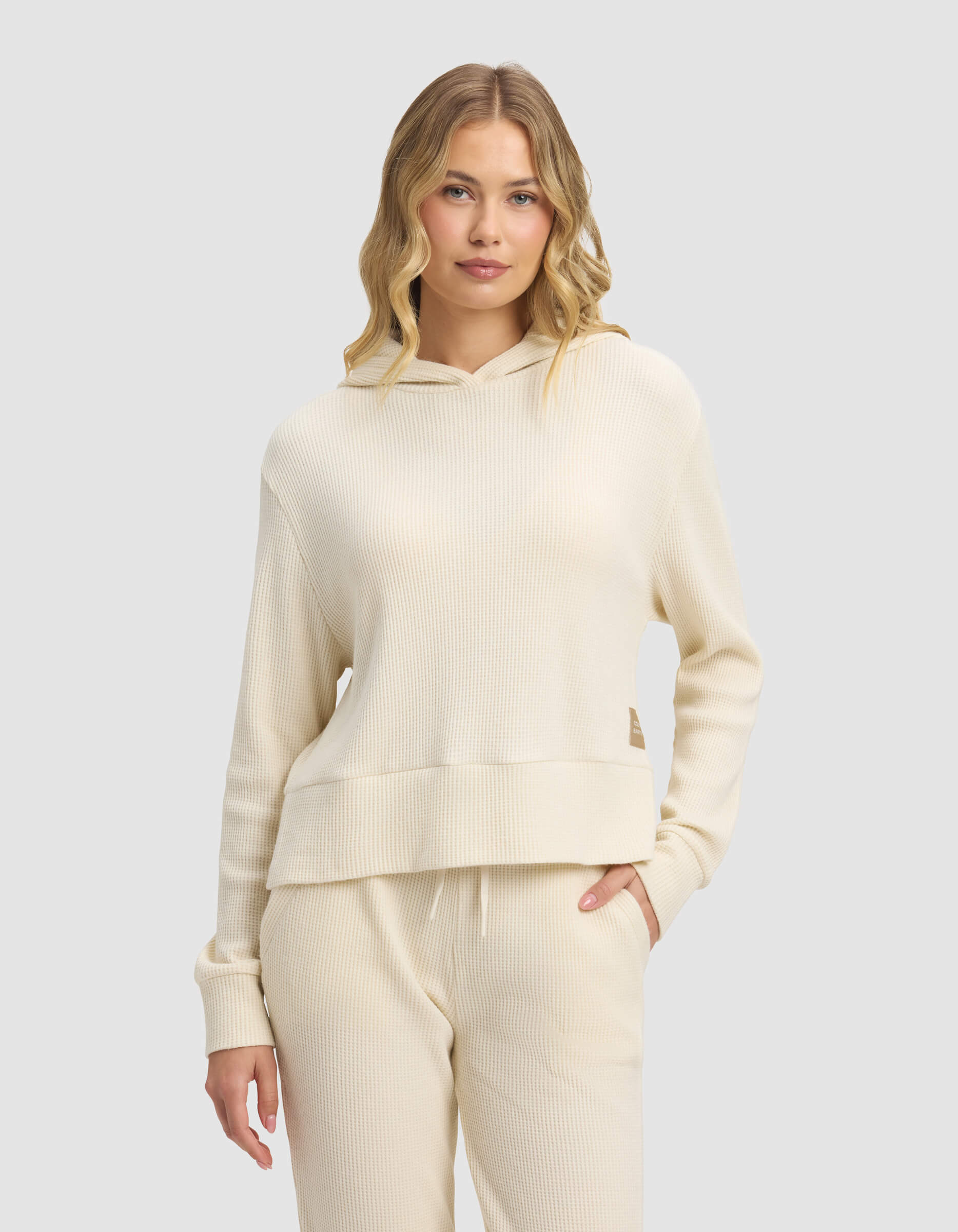 A woman with long wavy blonde hair models the Cozy Earth Women's Blake Waffle Hoodie in cream, standing against a light gray background with a relaxed and confident expression.