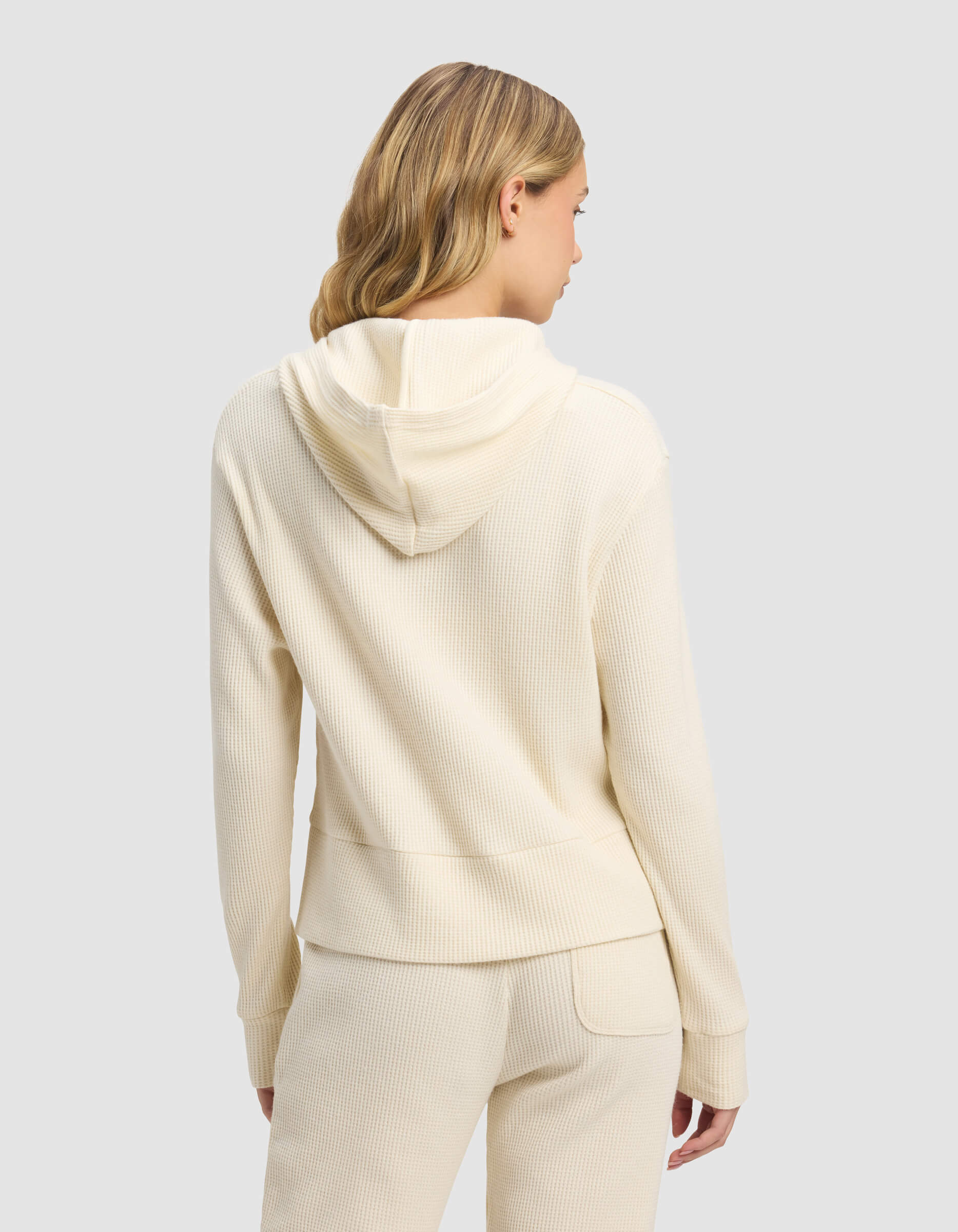 A woman with blonde hair is seen from the back, wearing the Cozy Earth Women's Blake Waffle Hoodie in cream and matching pants, set against a plain light gray background.|Color:Coconut