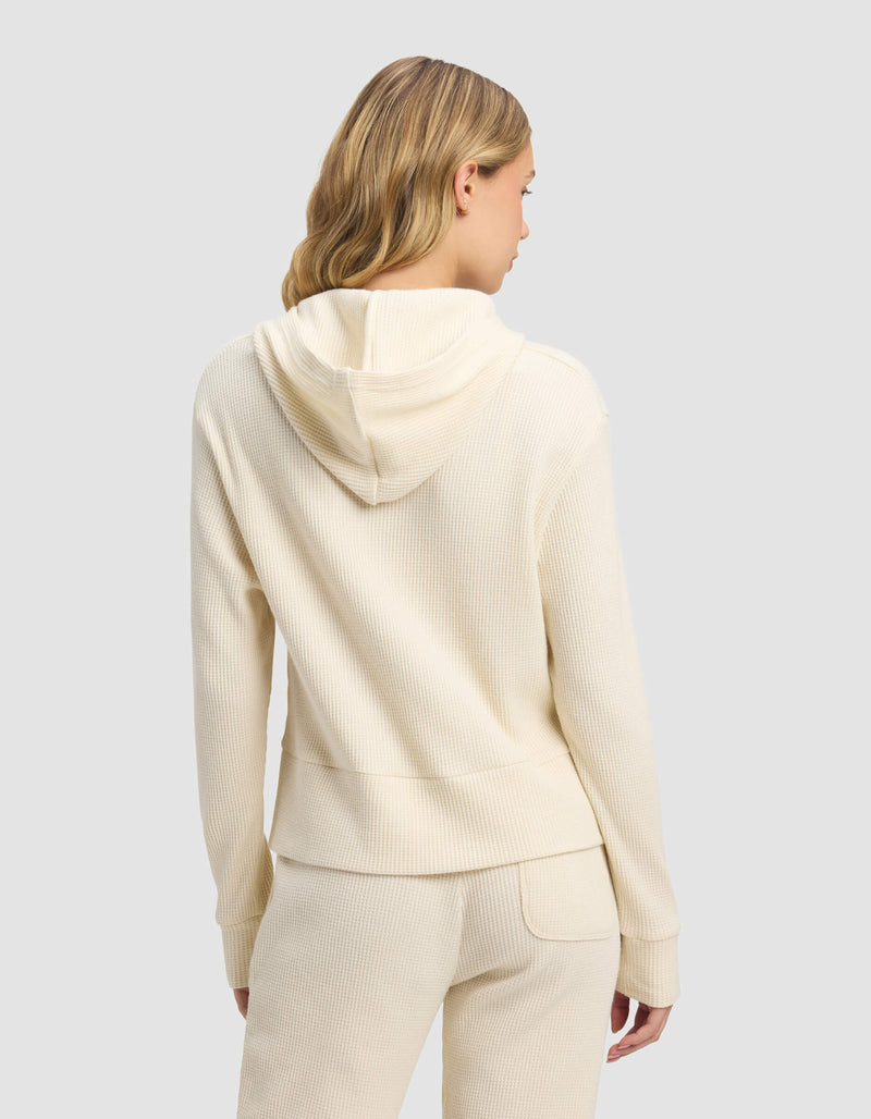 A woman with blonde hair is seen from the back, wearing the Cozy Earth Women's Blake Waffle Hoodie in cream and matching pants, set against a plain light gray background.