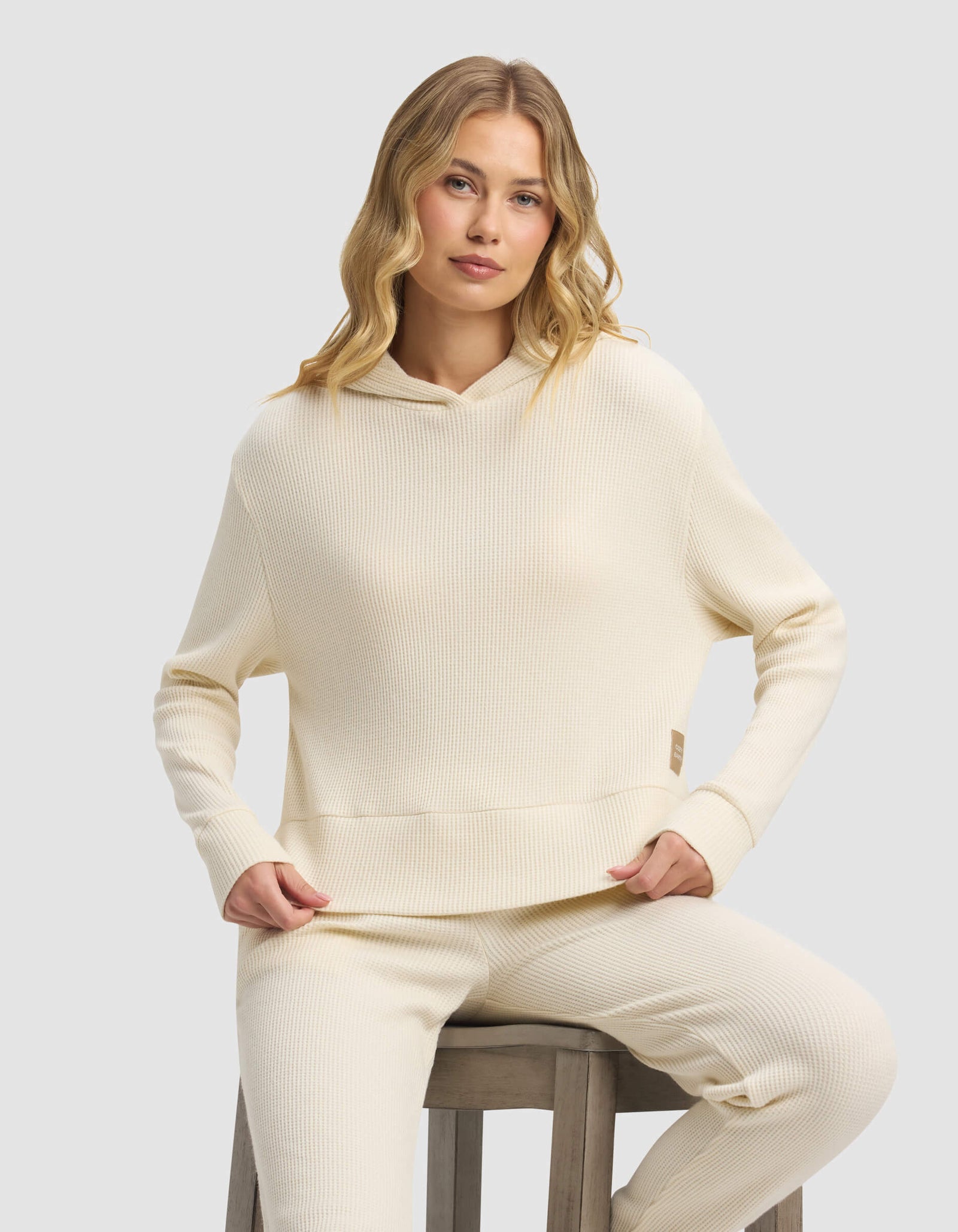 A woman with long, wavy blonde hair wears the Cozy Earth Women's Blake Waffle Hoodie in cream, paired with matching pants, while sitting on a wooden stool against a plain light gray background.