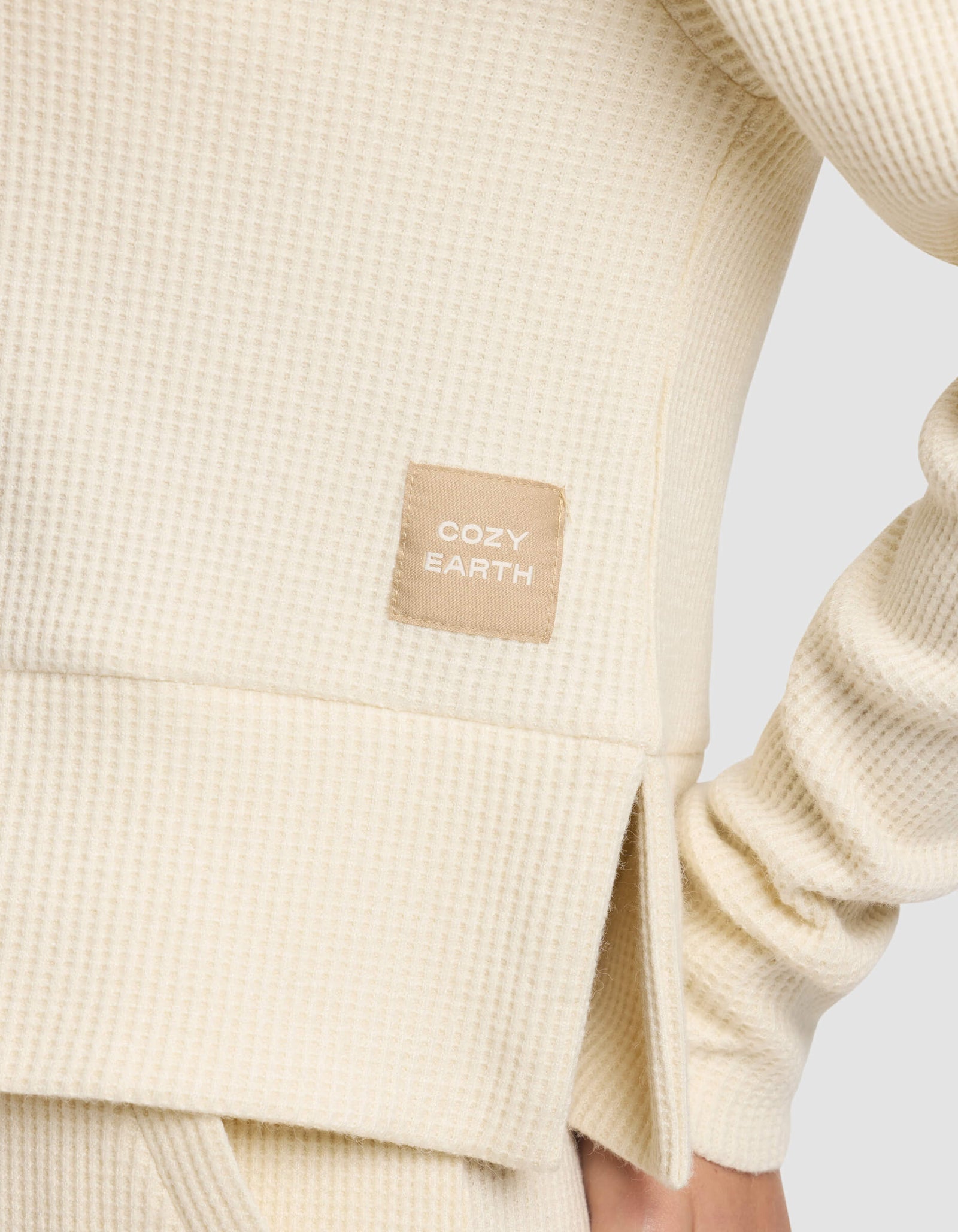 Close-up of the Women's Blake Waffle Hoodie by Cozy Earth in cream, showcasing its textured fabric and a stitched "COZY EARTH" tag. The wearer’s arm is bent at the elbow.