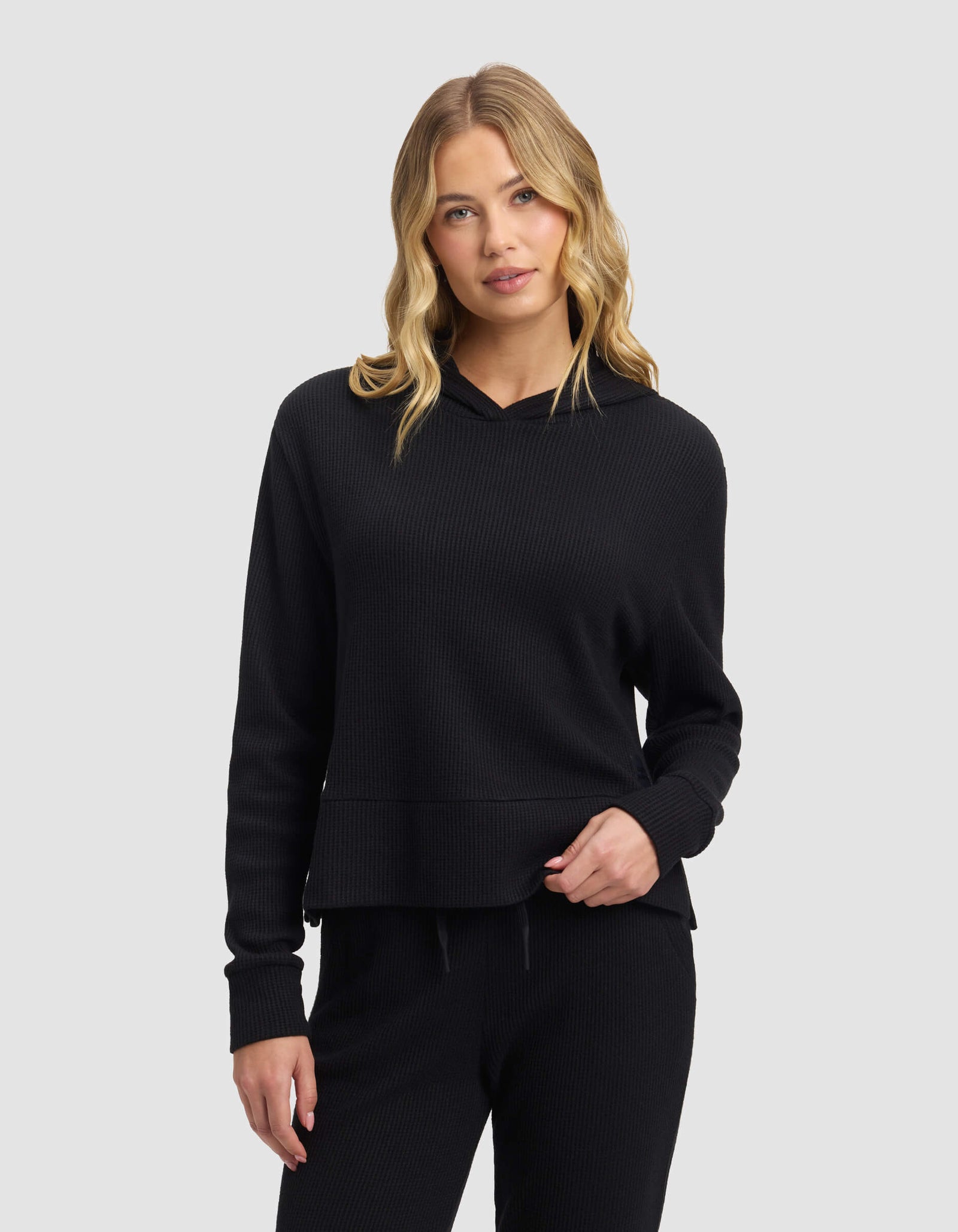 A woman with long, wavy blonde hair wears the Cozy Earth Women's Blake Waffle Hoodie in black, paired with matching black pants, standing against a plain light gray background.