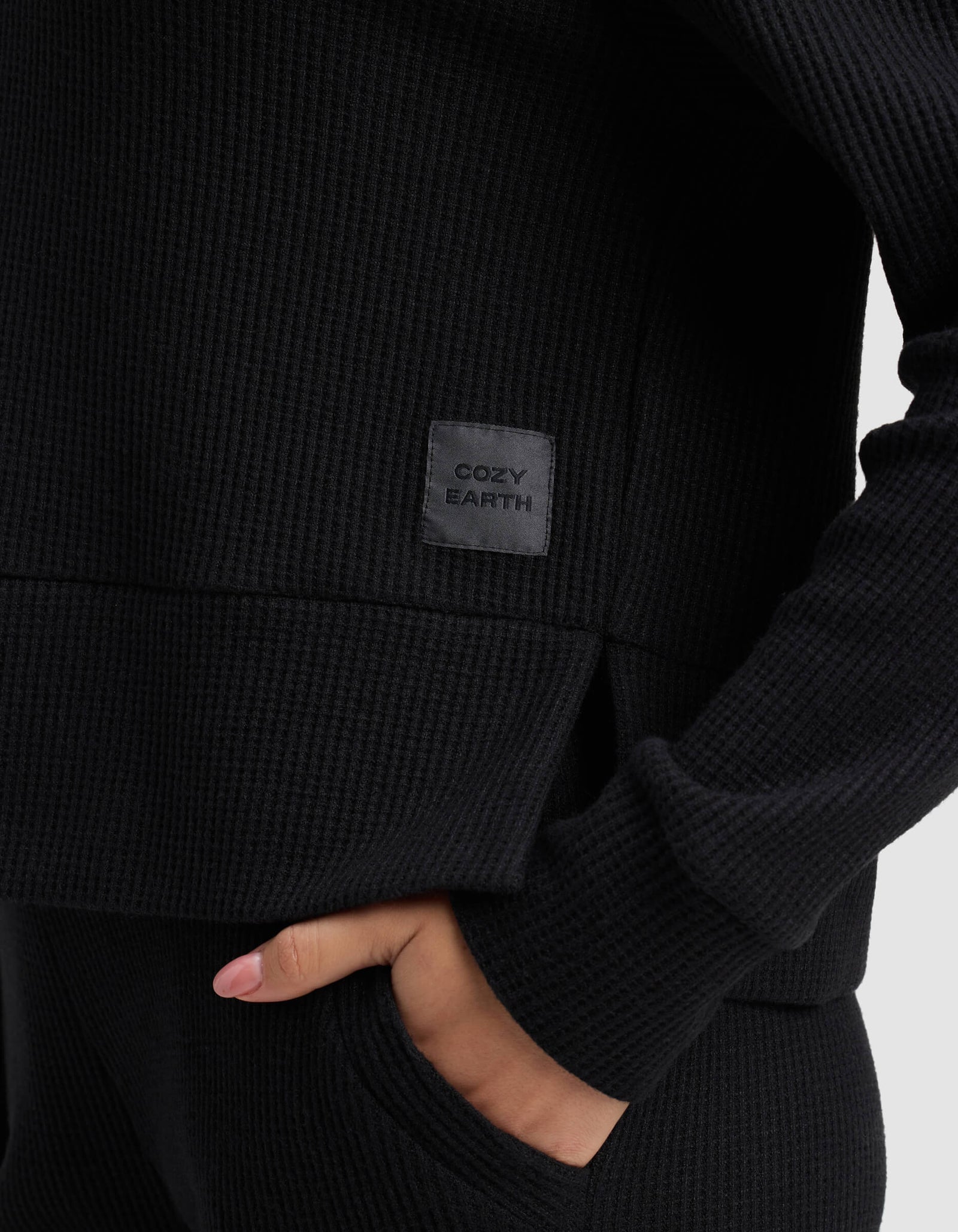 A woman wears the Cozy Earth Women's Blake Waffle Hoodie in black, hand in pocket; a gray patch on the shirt displays the brand name.