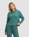 A woman with wavy blonde hair wears the Cozy Earth Women's Blake Waffle Hoodie in teal and matching pants, standing with one hand in her pocket against a plain light background.|Color:Sea Green