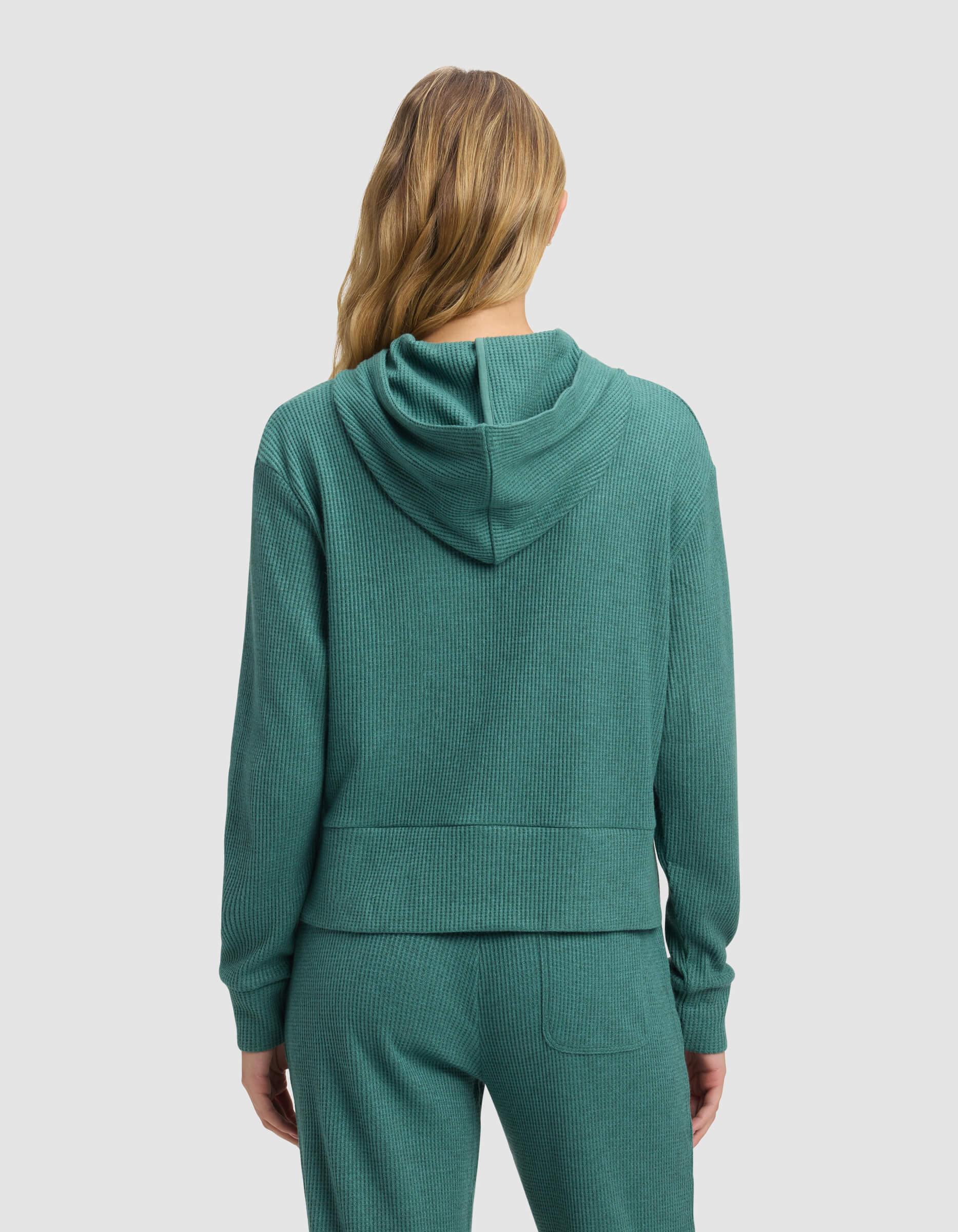 A woman with long, wavy, blonde hair is seen from the back wearing Cozy Earth's Women's Blake Waffle Hoodie in teal with matching pants against a plain, light background.
