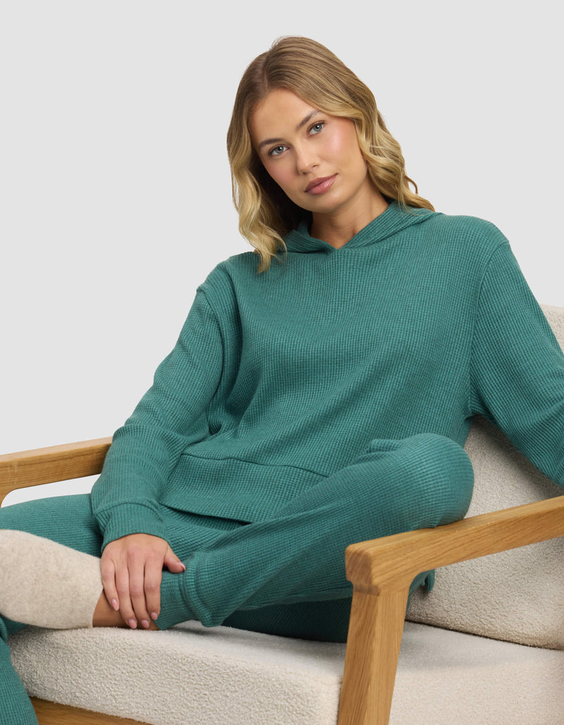 A blonde woman sits relaxed on a beige armchair, wearing the Cozy Earth Women's Blake Waffle Hoodie in teal, with one arm resting on the chair and her legs crossed.