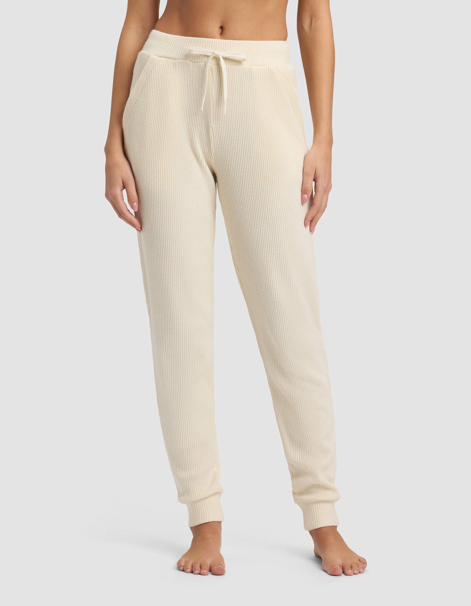 A person wears Cozy Earth's Women's Blake Waffle Jogger in cream, featuring a ribbed knit, drawstring waist, and pockets, while standing barefoot against a plain background.