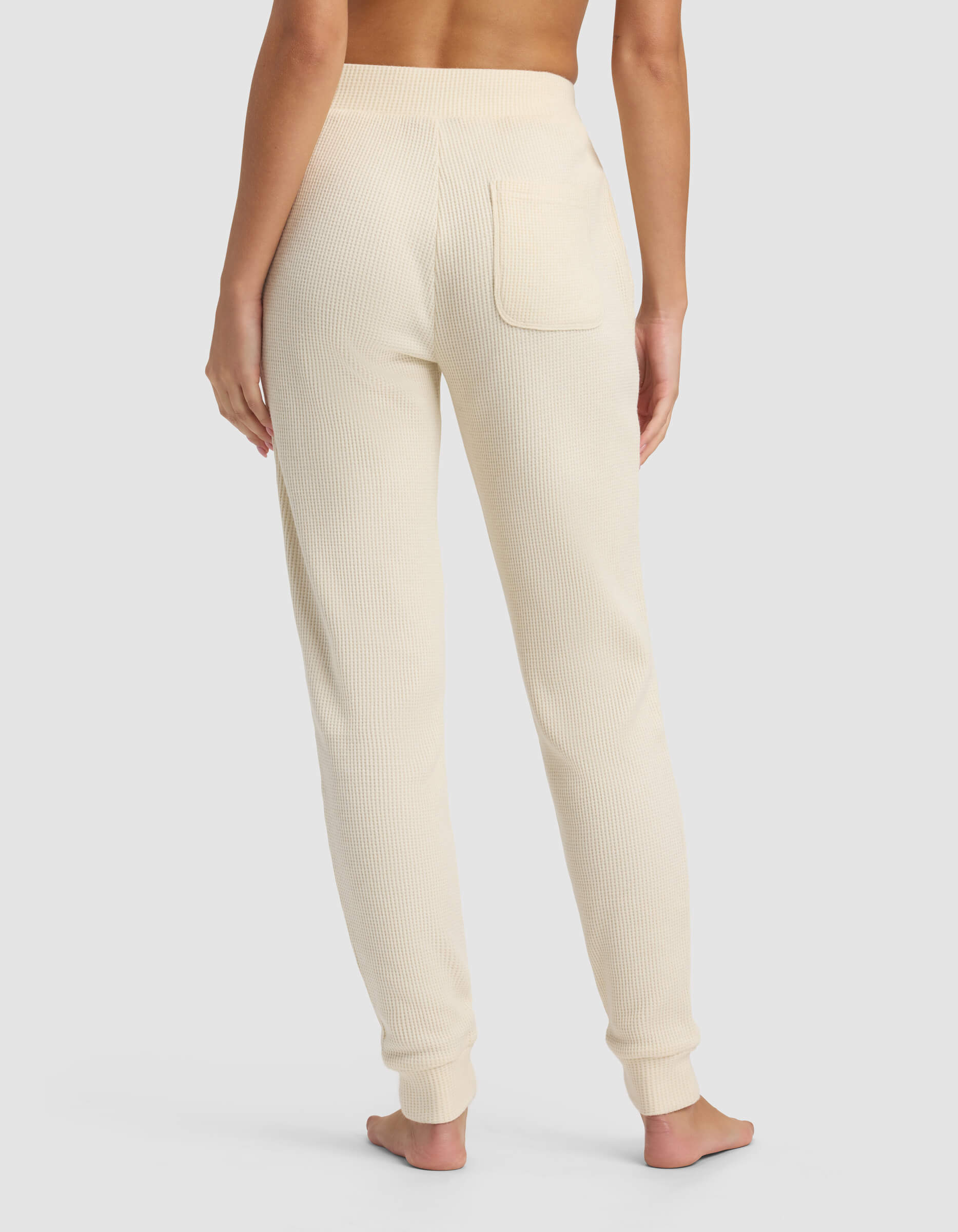 A person stands barefoot facing away, wearing Cozy Earth's Women's Blake Waffle Jogger in a cream high-waisted ribbed knit with a single back pocket, set against a plain light gray background.