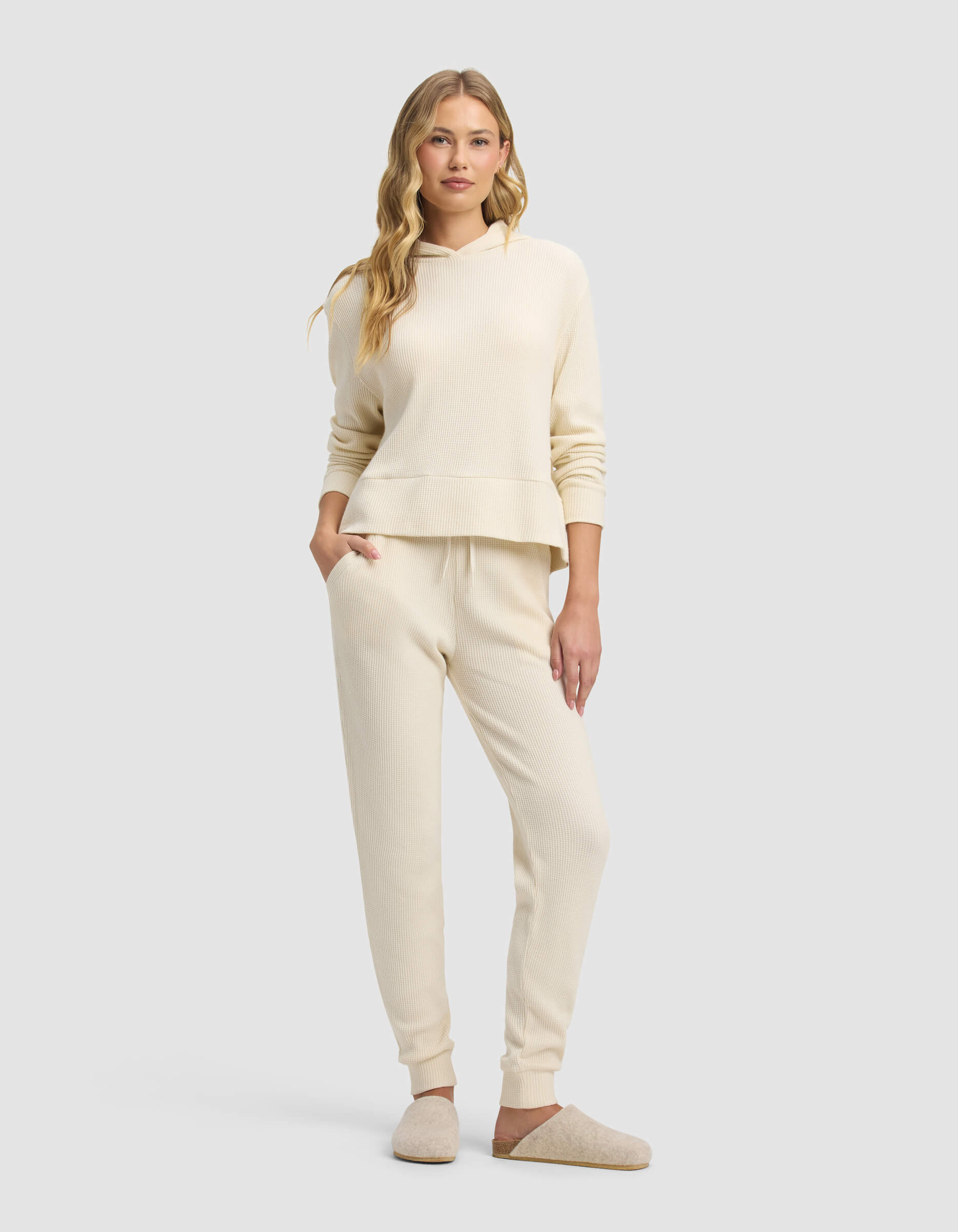 A woman with long wavy blonde hair stands against a plain background, wearing Cozy Earth's cream Women's Blake Waffle Jogger and matching hooded sweatshirt, paired with beige slippers. One hand rests in her pocket.